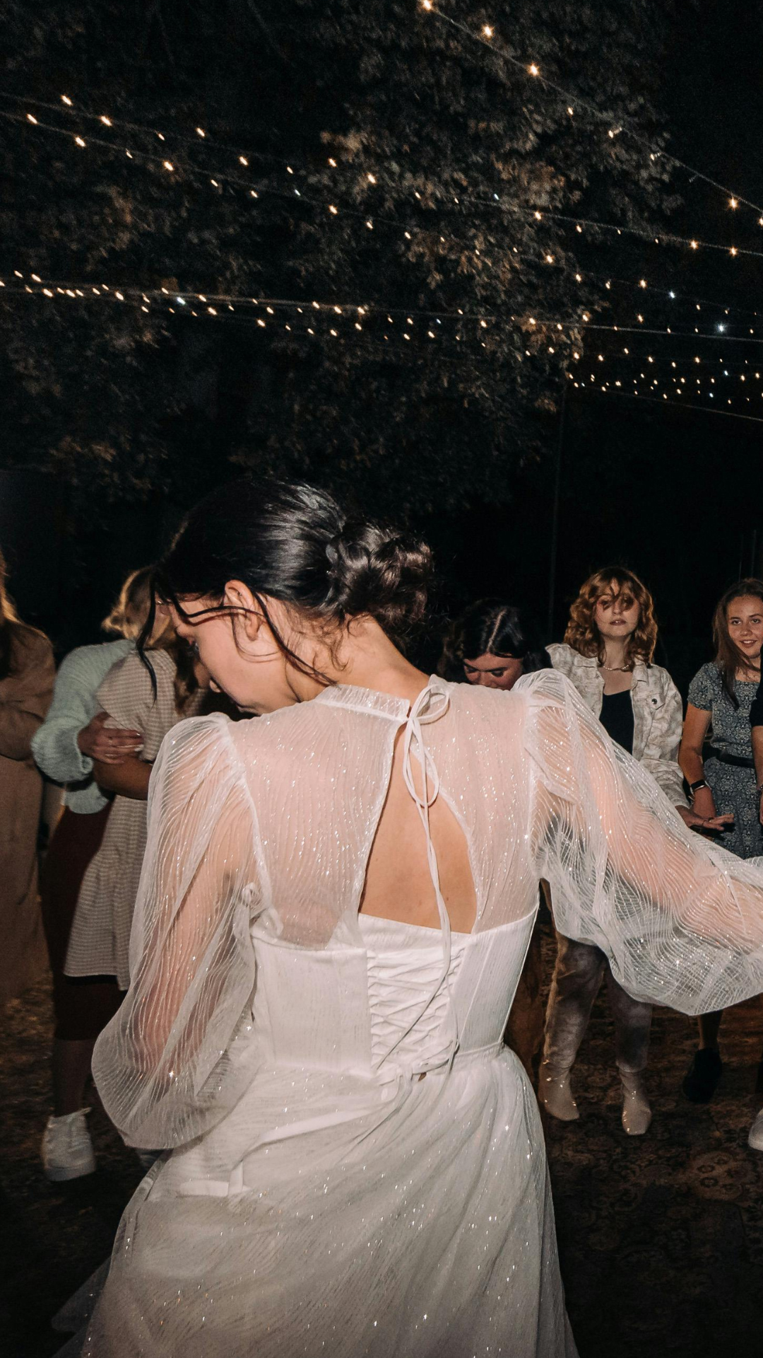 A woman in a white dress with a ribbon tie at the back, dancing at an outdoor event with string lights, surrounded by other women.