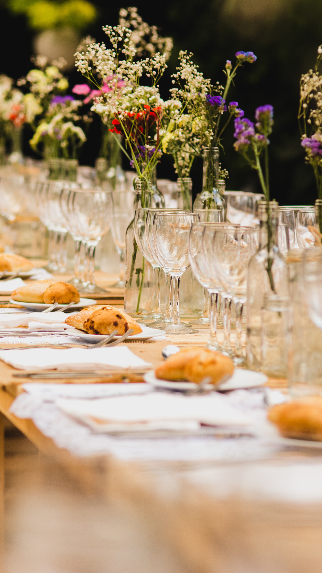 A table setting with wine glasses, bottles with flowers, plates, and pastries outdoors.