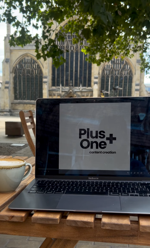 A laptop on a wooden table displaying the words 'Plus One+ content creation' with a cappuccino beside it. In the background, a historic church with large arched stained glass windows is visible, partly obscured by some tree leaves.