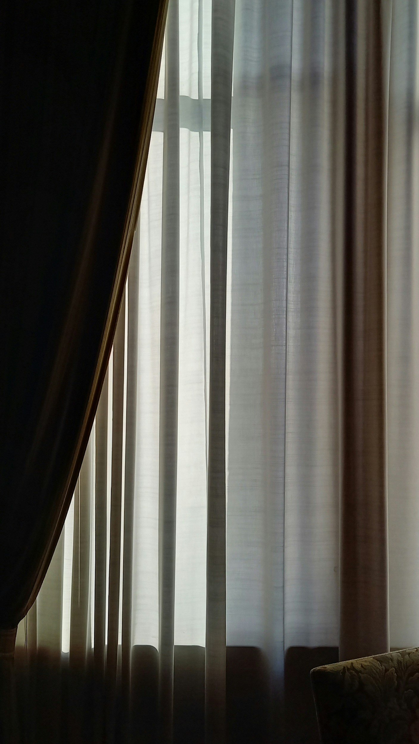 Curtains with sunlight coming through, partially opened window behind them, partial view of a dark furniture piece on the left and a patterned armrest on the right.