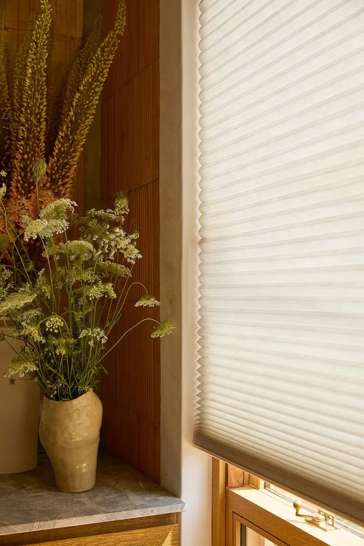 A beige vase with white and yellow-green flowers and tall grass-like plants on a stone surface next to a window with a white pleated shade.