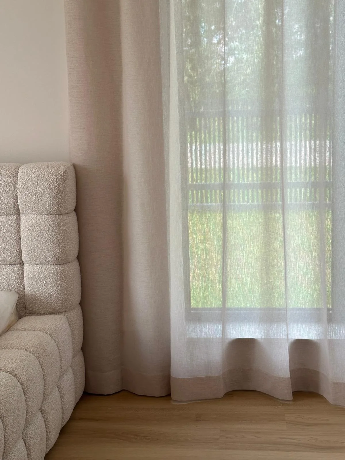 A beige upholstered headboard with a textured pattern is on the left side in front of a light-colored wall. To the right, there are floor-length sheer white curtains covering a window, through which a balcony railing and greenery outside are visible.