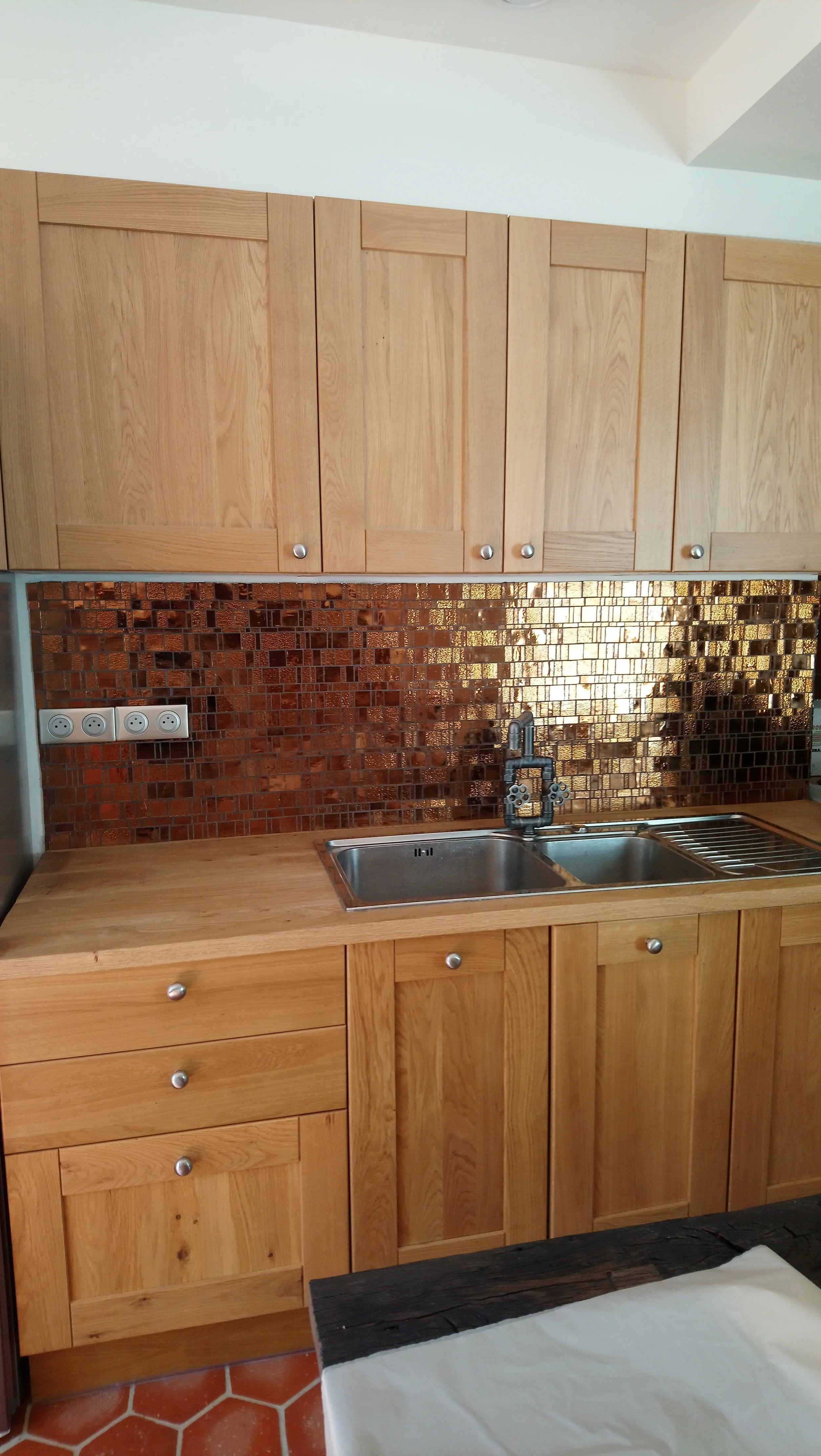 Cuisine avec armoires en bois clair, évier double en acier inoxydable, plan de travail en bois, mur en mosaïque de carreaux cuivrés, prises électriques blanches, table en bois avec nappe blanche et une pièce en noir à l'avant.