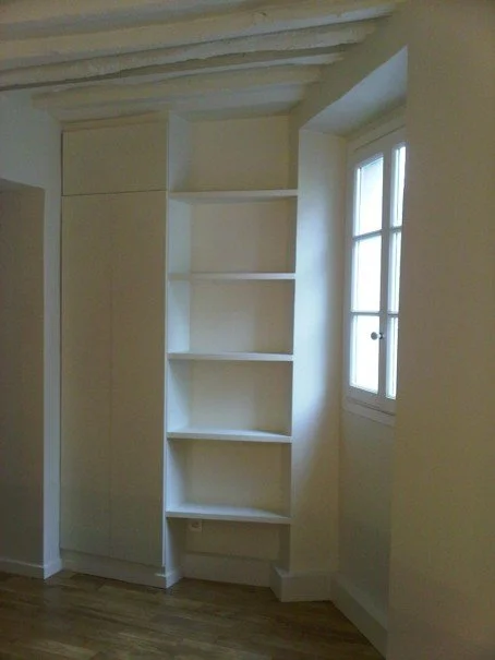 Une pièce avec un mur blanc, une étagère blanche à quatre niveaux, une fenêtre avec volets en bois, et un plafond avec des poutres apparentes.