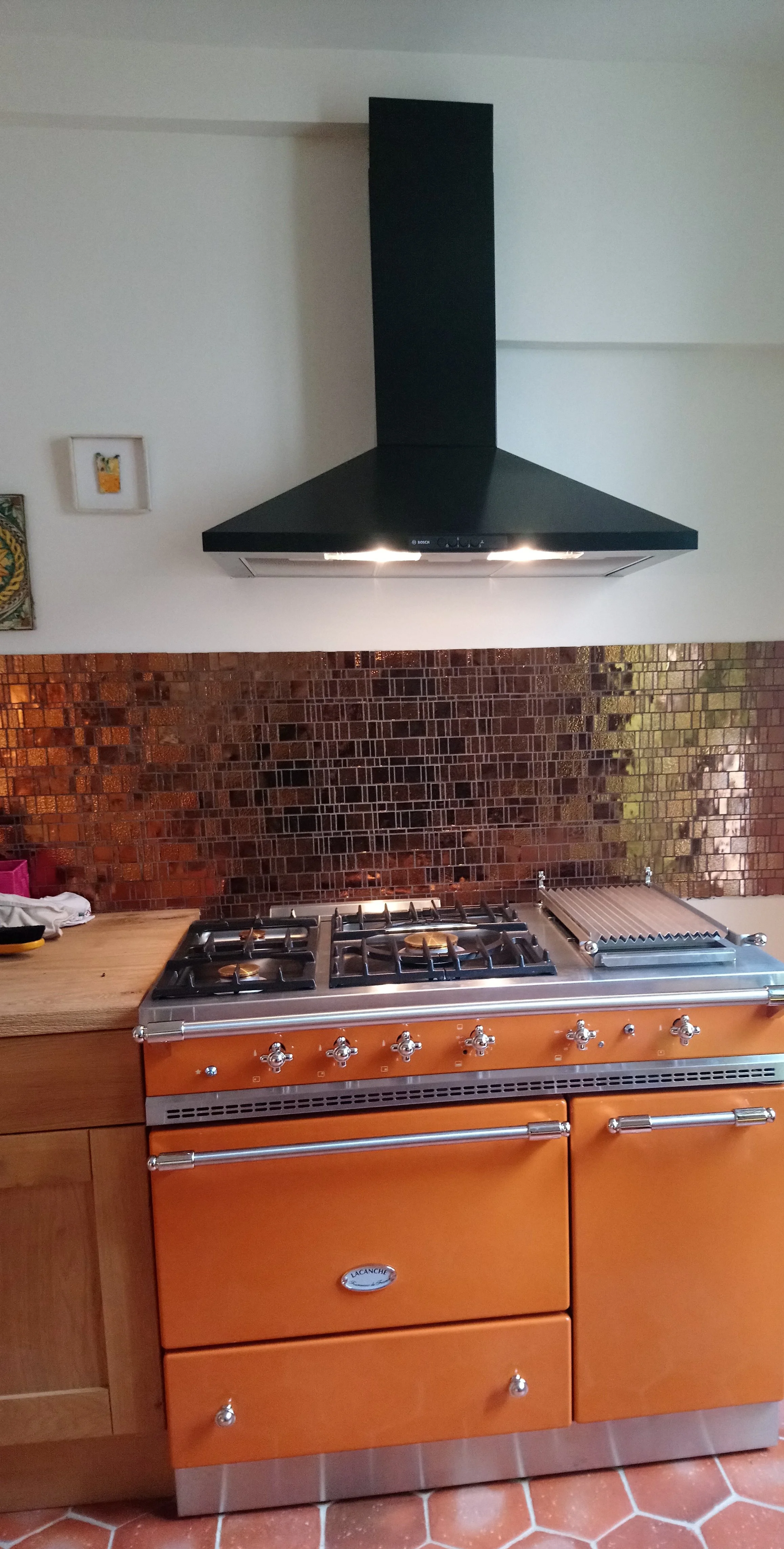 Cuisinière à gaz en bois orange avec six boutons, deux tiroirs en dessous, plan de travail en bois à gauche, backsplash en mosaïque de petits carreaux en bronze et noir, hotte de cuisson noire au mur, partie du sol en carreaux hexagonaux orange.