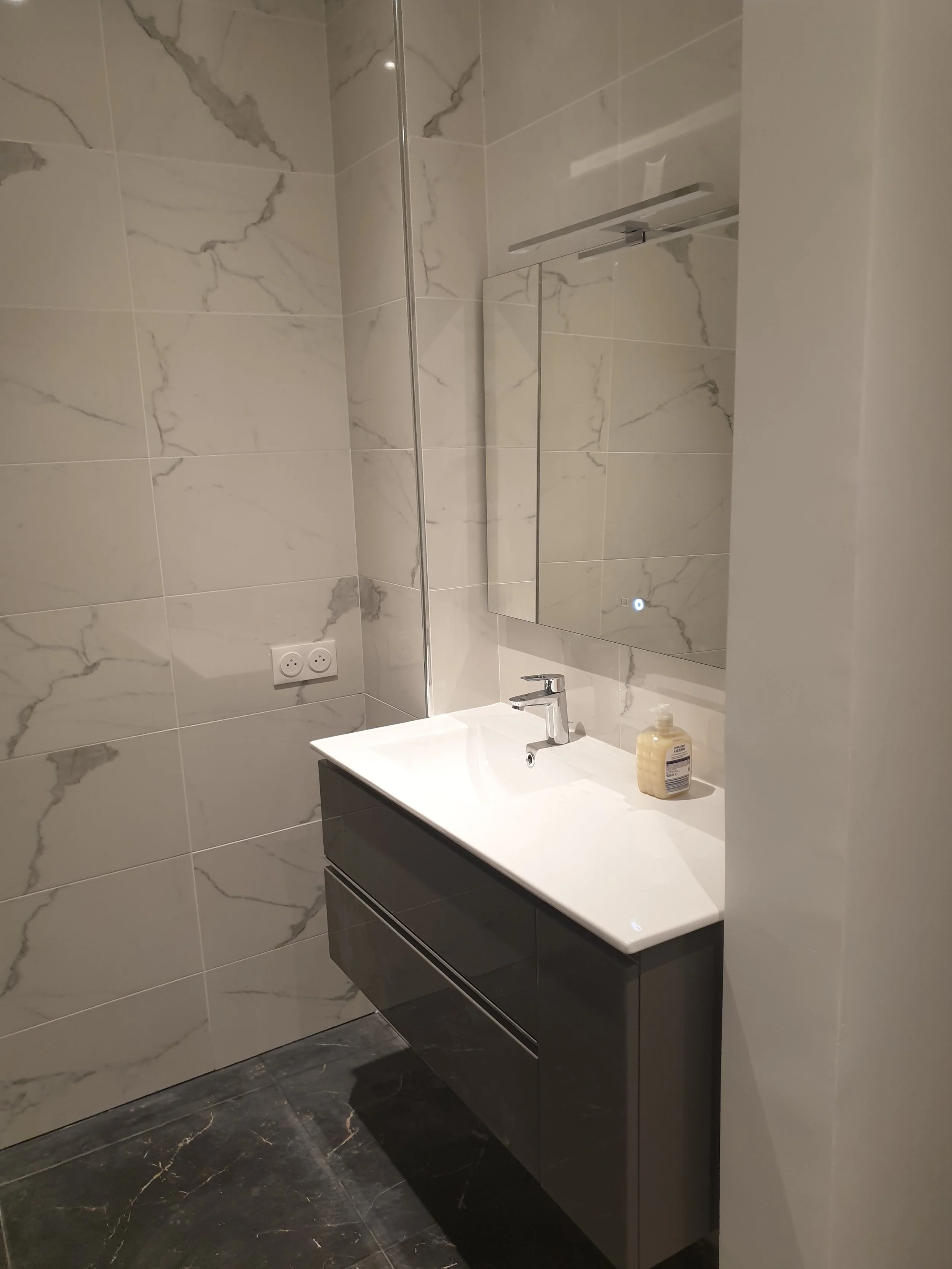Un miroir au-dessus d'un lavabo dans une salle de bain moderne avec murs en carreaux de marbre blanc et sols en marbre noir.