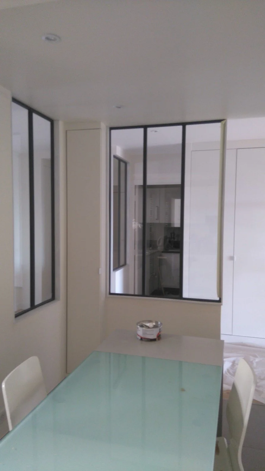 Intérieur d'une pièce moderne avec un mur en verre noir, une table en verre avec des chaises blanches, et une cuisine visible à l'arrière.