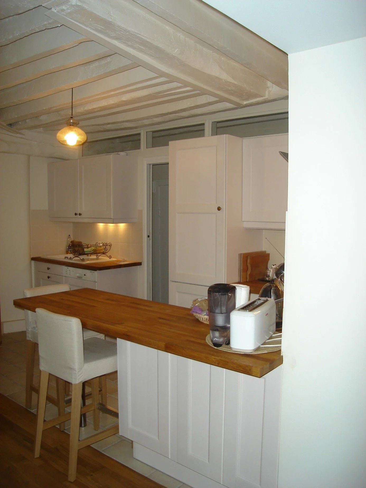 Cuisine avec table en bois, deux chaises en tissu blanc, cuisine blanche avec des armoires, appareils électroménagers, et un plafond en bois apparent avec une lumière