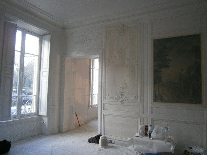 Salle élégante avec murs ornés de moulures et un tableau, grande fenêtre laissant entrer la lumière, mobilier minimal, quelques objets sur une petite table