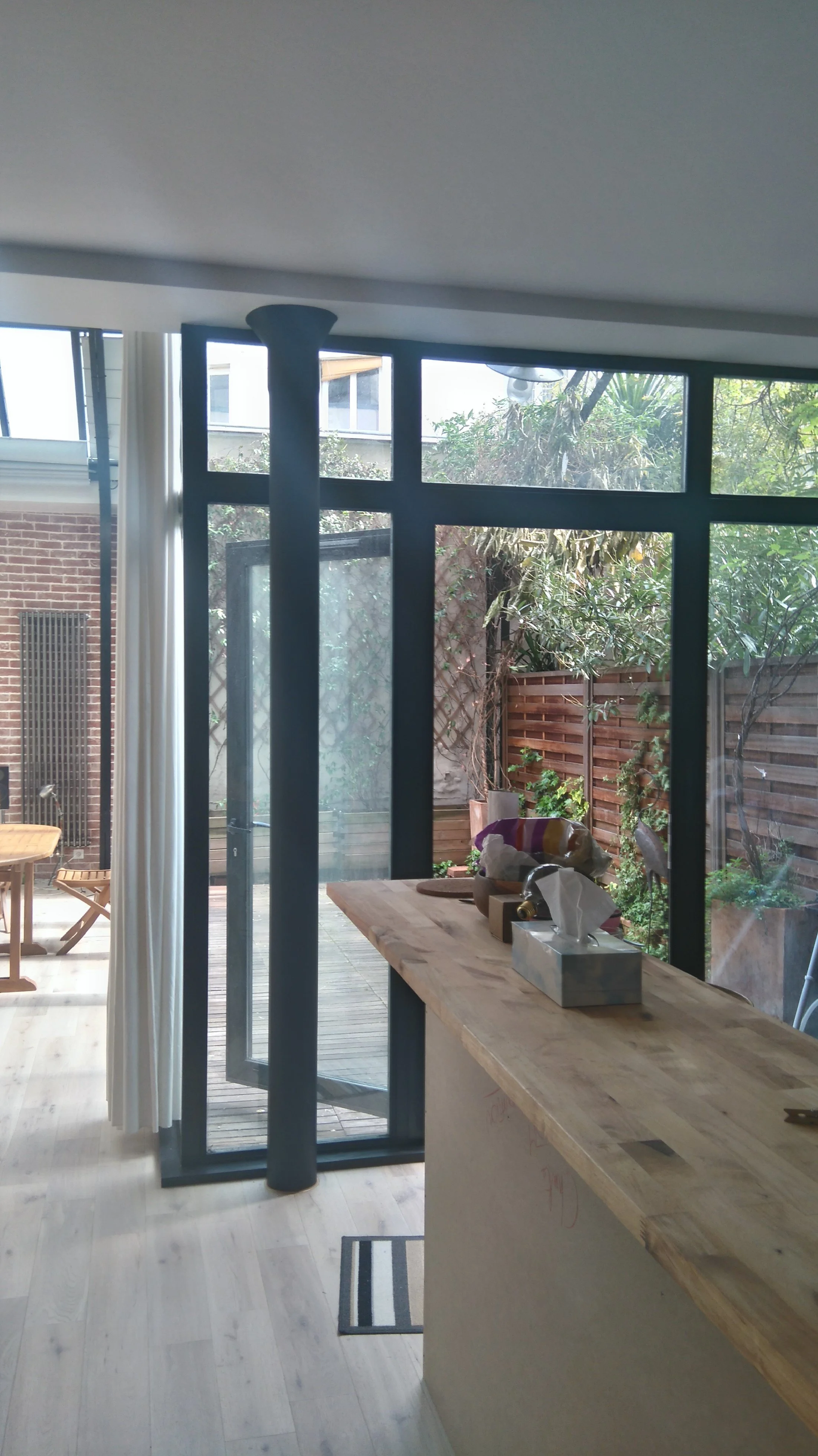 Vue intérieure d'une pièce avec une grande baie vitrée donnant sur un jardin avec des plantes et une clôture en bois, un comptoir en bois avec des objets dessus, un tapis noir et blanc, et un tapis gris dehors.