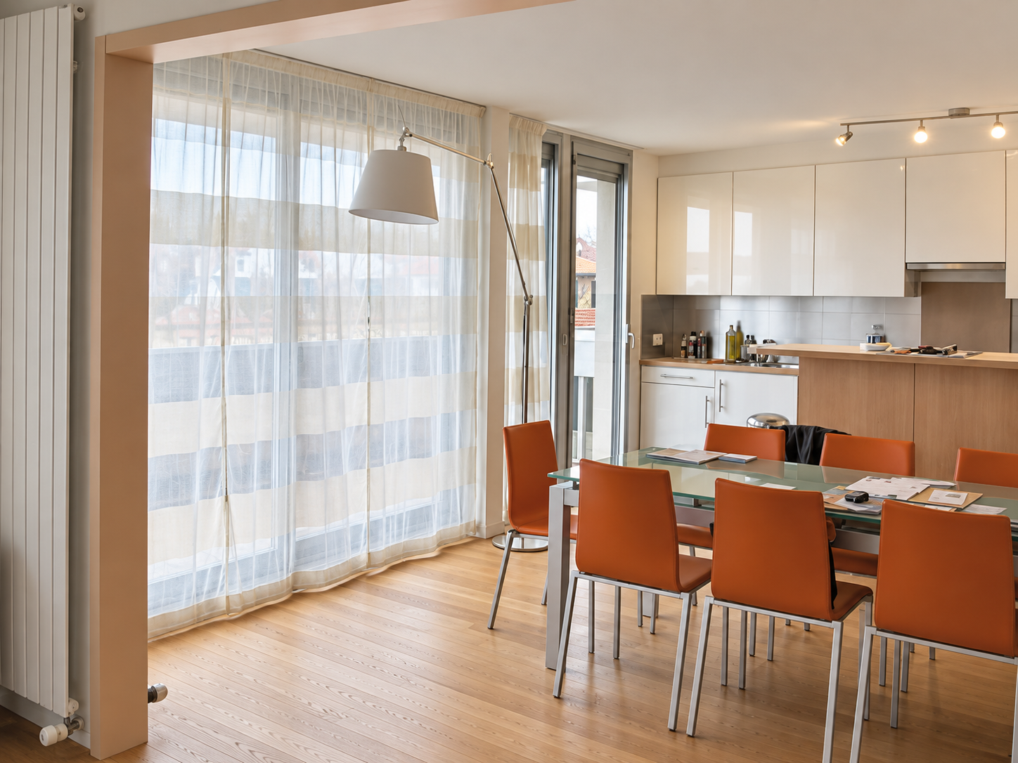 Salon de repas moderne avec une table en verre et des fauteuils orange, cuisine en arrière-plan, grande fenêtre avec rideaux transparents, parquet en bois clair.