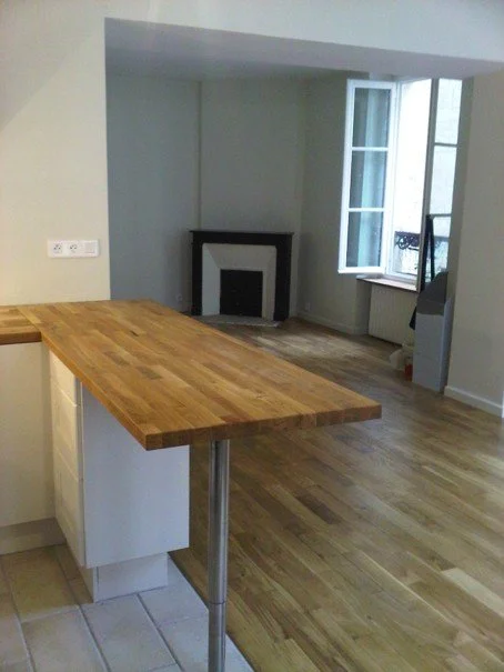 Salon avec une fenêtre ouverte, un carrelage au sol près d'une cuisine avec un plan de travail en bois, un coin cheminée et un radiateur