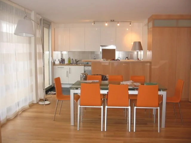 Salle à manger avec table blanche, huit chaises orange, cuisine en arrière-plan, sol en bois, grande fenêtre avec voilage blanc et lampadaire à gauche.