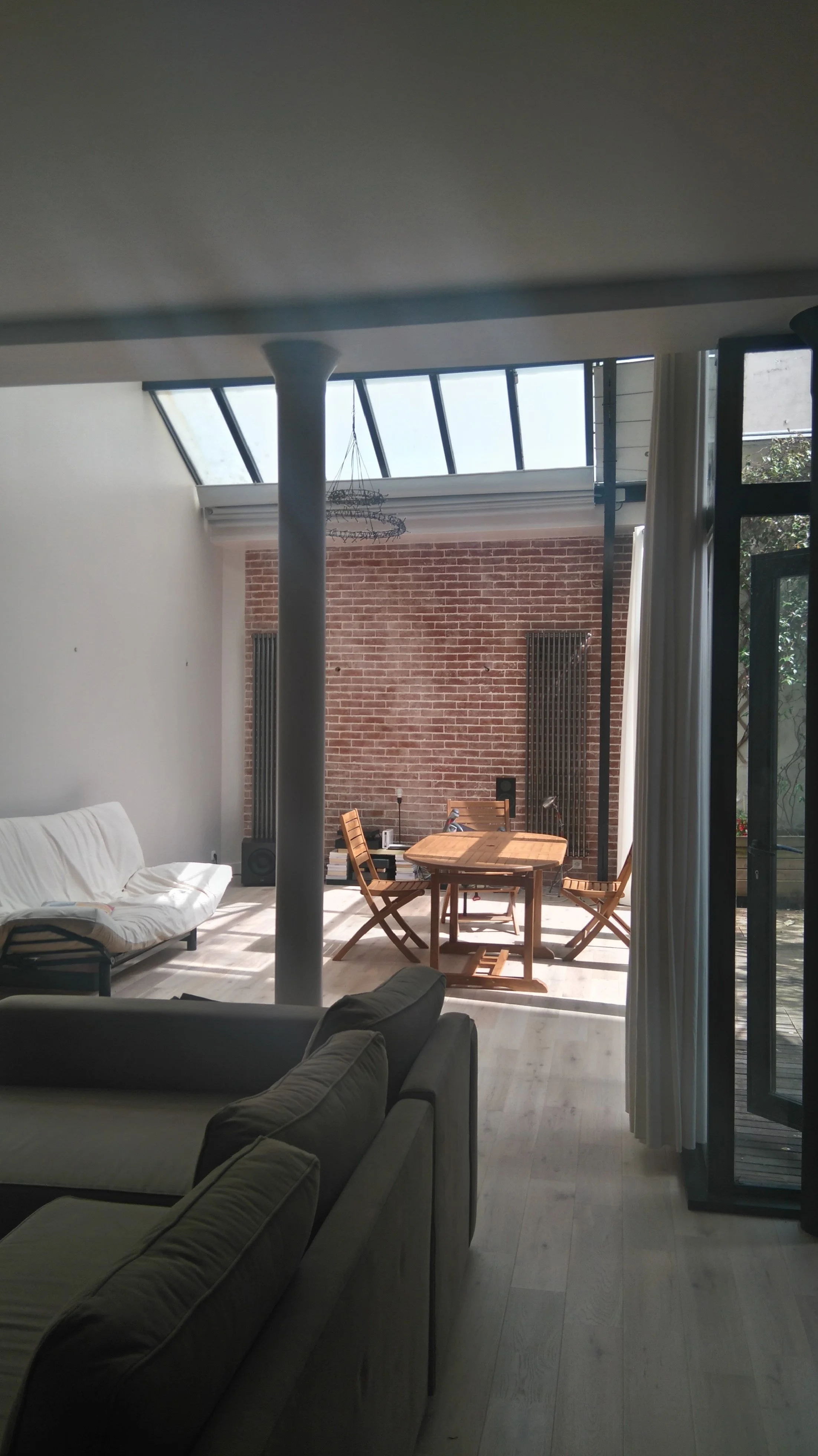 Intérieur d'un salon avec un canapé gris, un espace salle à manger avec une table en bois et des chaises en bois, mur en briques rouges, grande fenêtre en verre avec rideaux blancs, et un plafond avec une structure en acier et un chandelier suspendu.