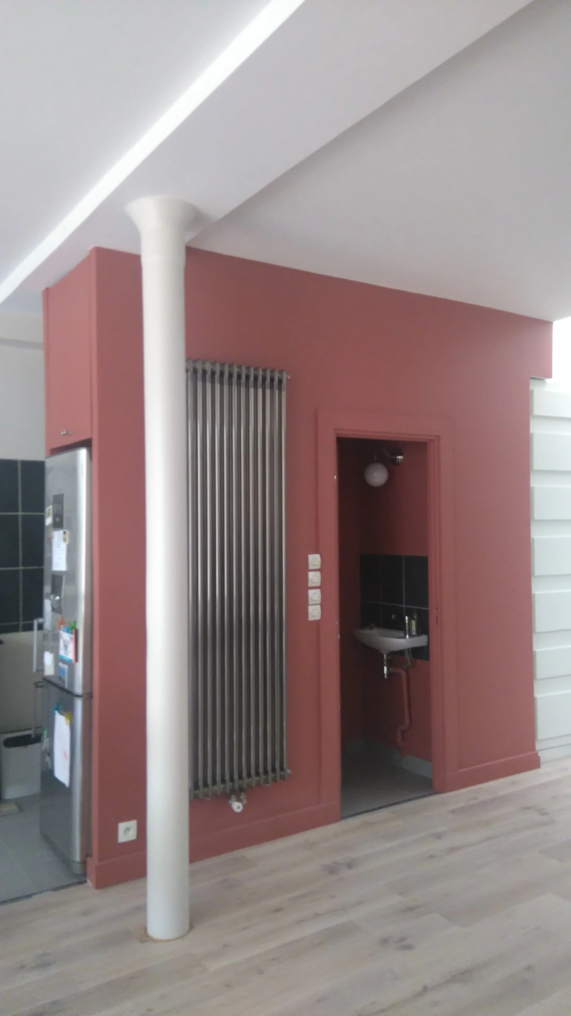Intérieur moderne avec mur rose et poutre blanche, radiateur vertical en métal, porte donnant sur un petit lavabo avec éclairage, sol en bois clair.