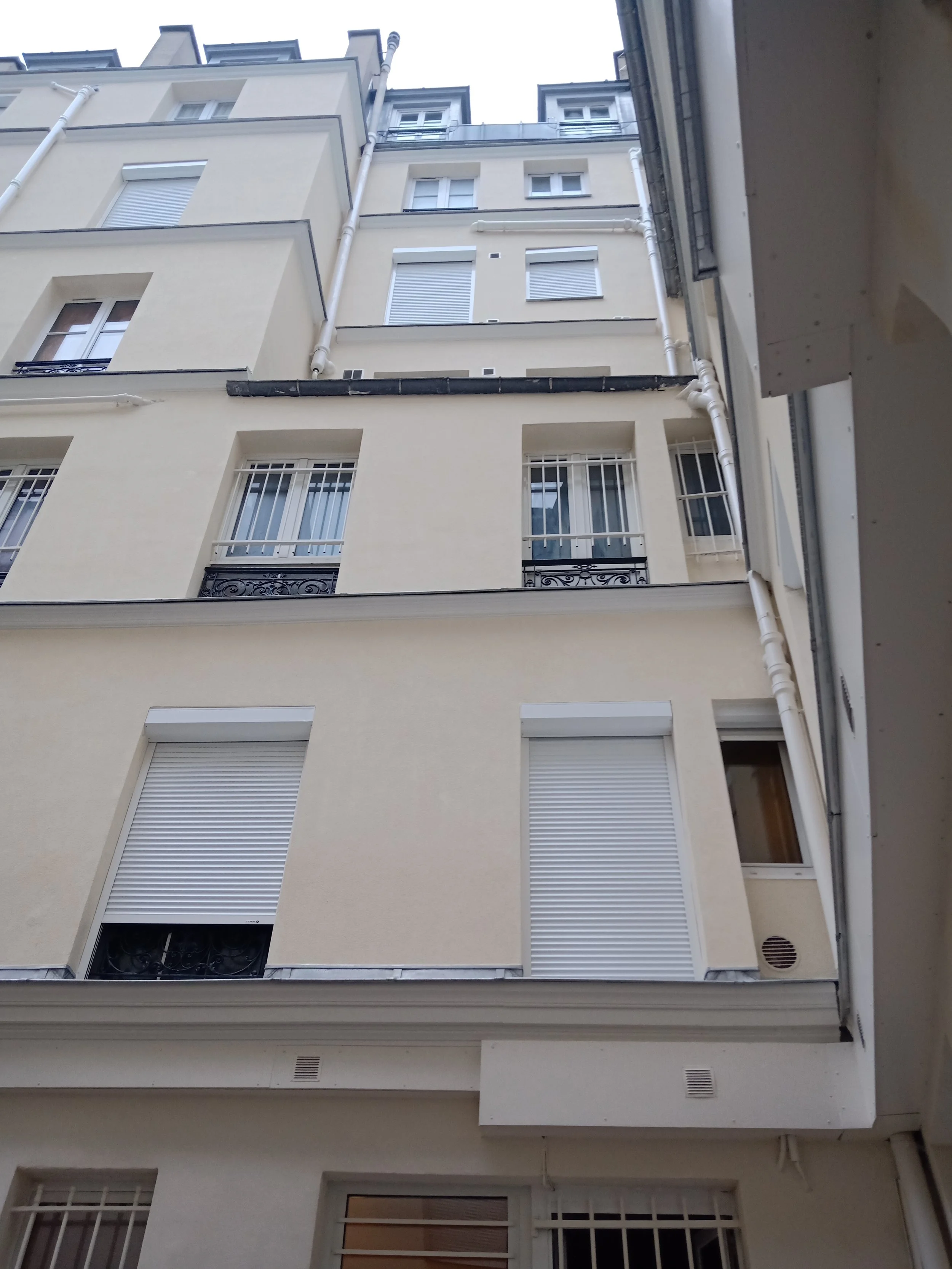 Façade d'un immeuble résidentiel avec plusieurs fenêtres, quelques unes équipées de volets roulants, et des balustrades en fer forgé. La photo est prise en contre-plongée depuis un petit espace entre deux bâtiments.