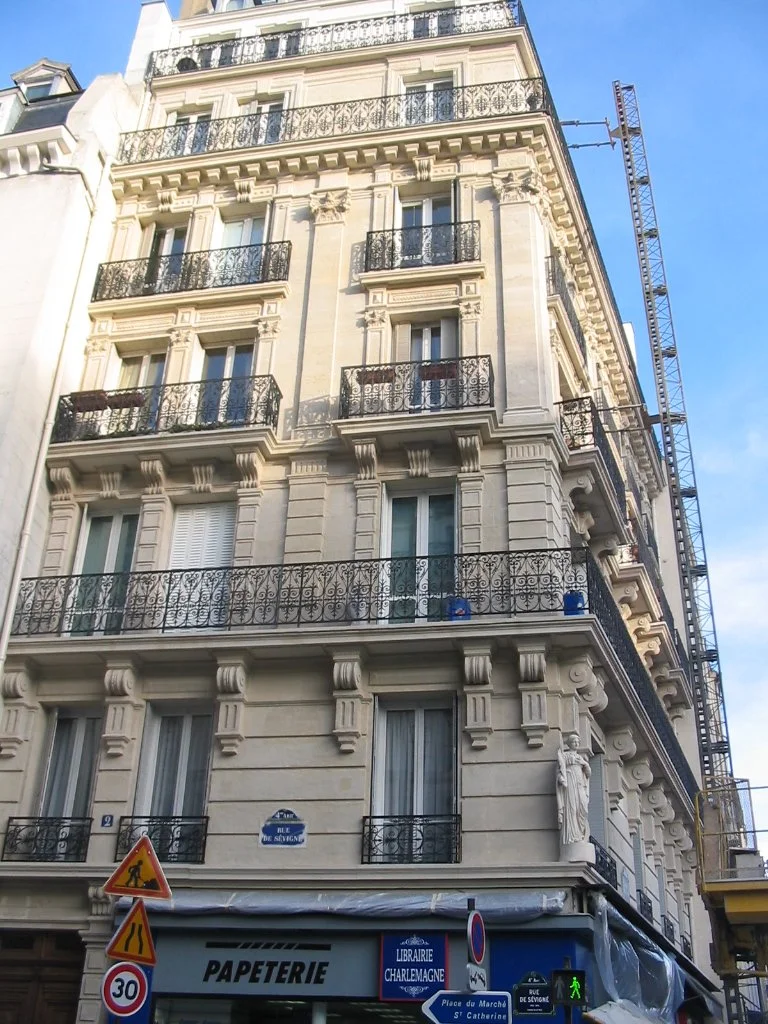 Immeuble haussmannien avec balcons en fer forgé, statues décoratives, et un bâtiment de papeterie au rez-de-chaussée, situé sur la rue de Sévigné à Paris, France.
