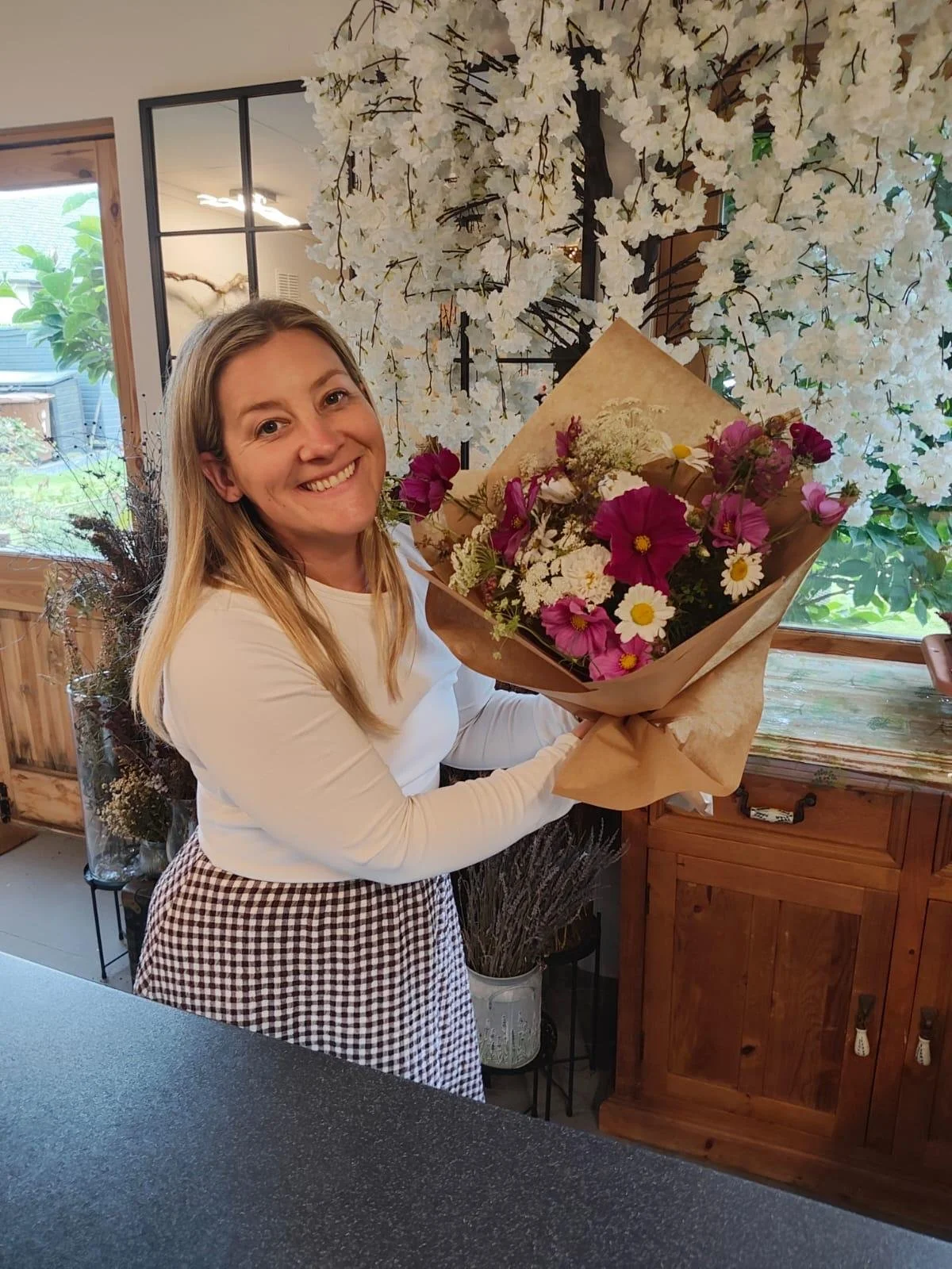 Smiling woman holding a colorful bouquet of fresh flowers in a flower shop decorated with white blossoms.