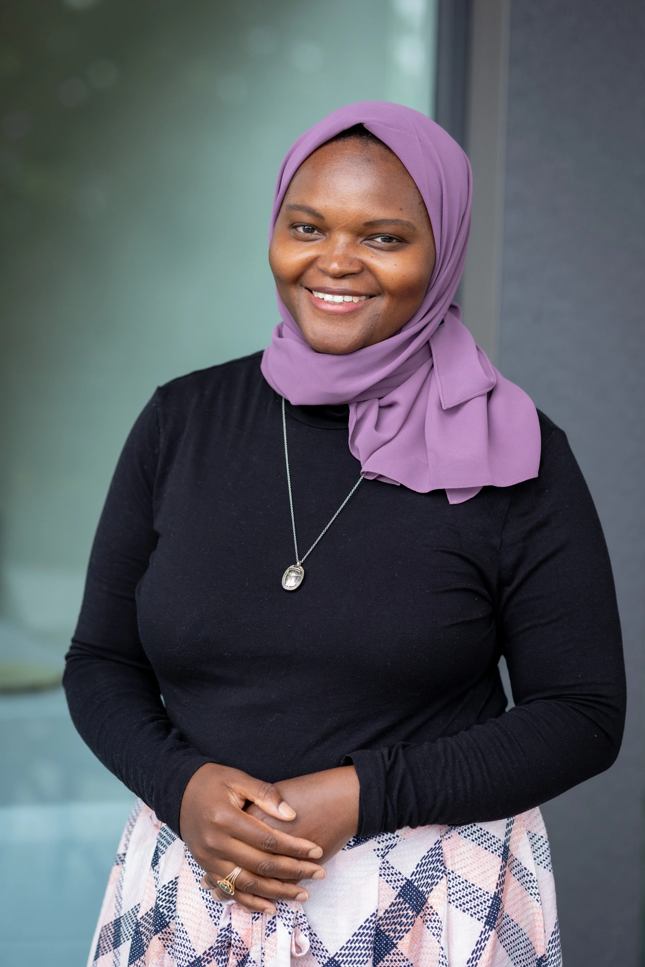 A woman smiling wearing a purple headscarf, black top, and patterned skirt.