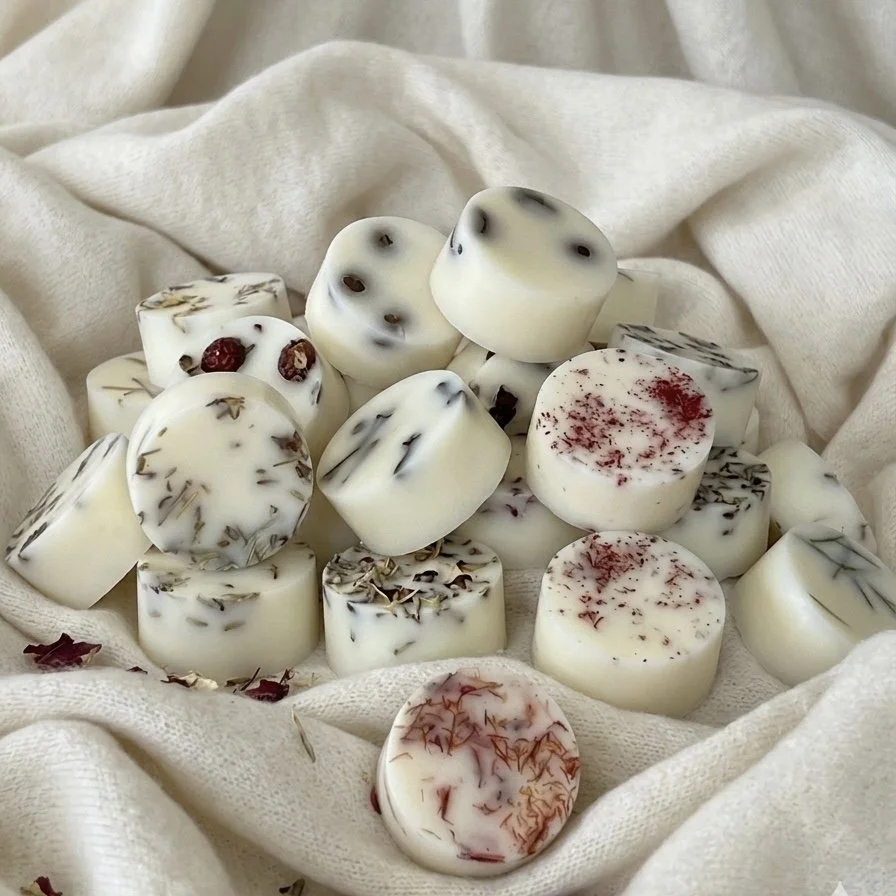 Several white scented candles with dried herbs and flowers embedded, placed on a soft, cream-colored fabric.