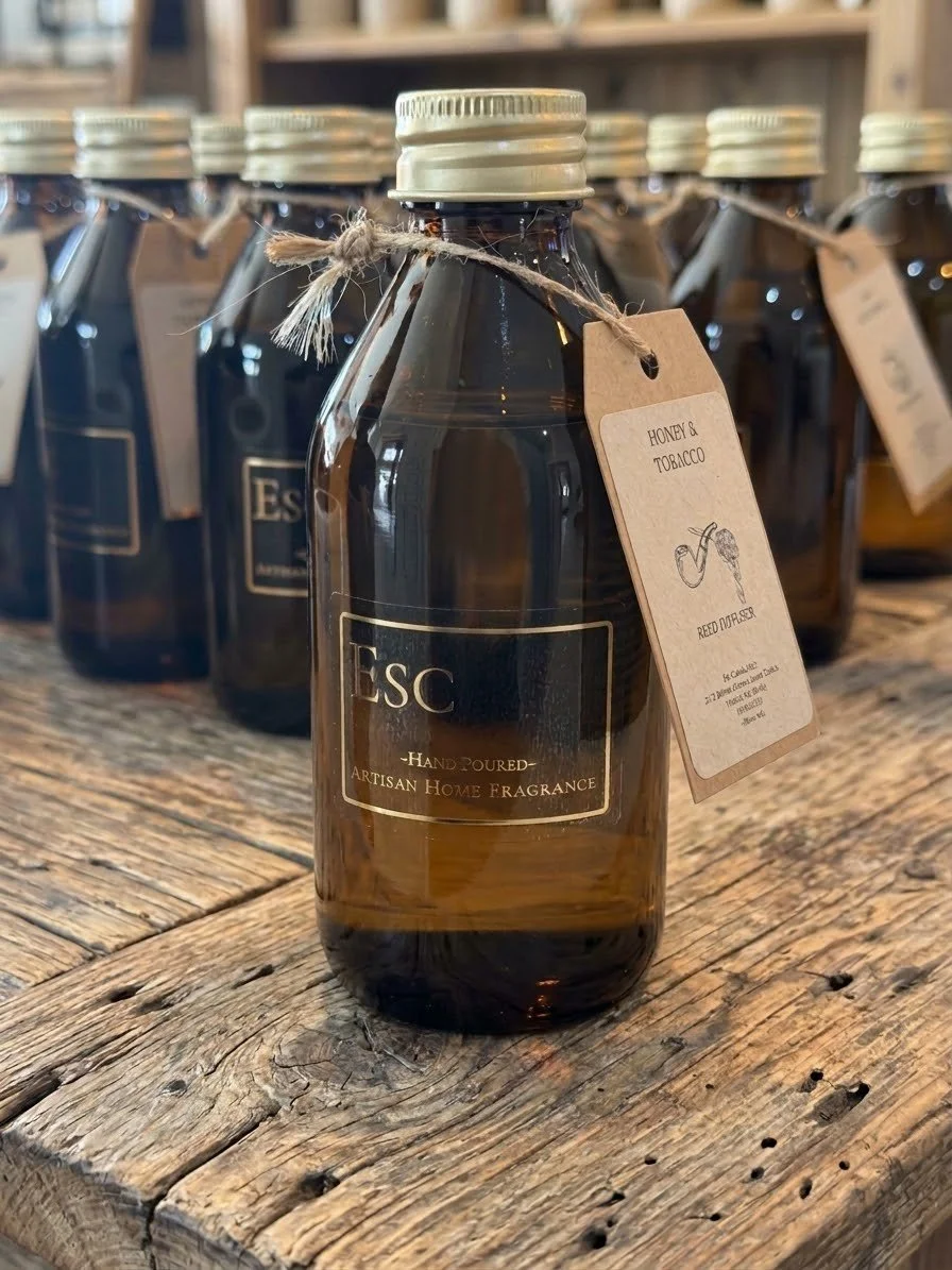 A dark brown glass bottle with a silver cap, labeled 'ESC Hand-Poured Artisan Home Fragrance,' with a tag that reads 'HONEY & TOBACCO' and an illustration of a pipe, placed on a rustic wooden surface.