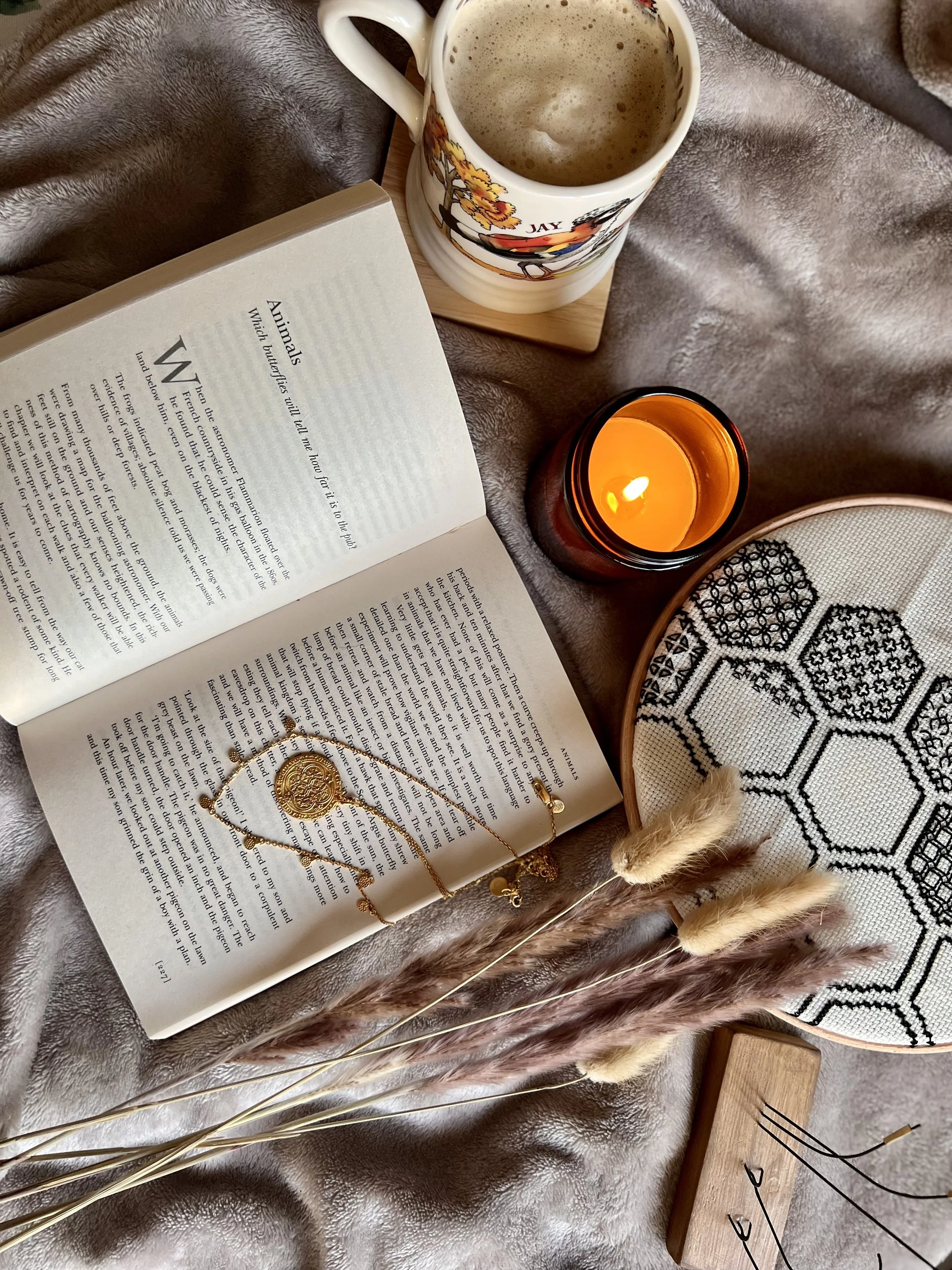 An open book with printed text on animals, a gold jewelry piece resting on it, a ceramic mug with a winter scene and the word 'JAY' on it, a lit yellow candle, dried decorative grasses, and a woven cloth with geometric patterns, all placed on a plush, brown fabric surface.