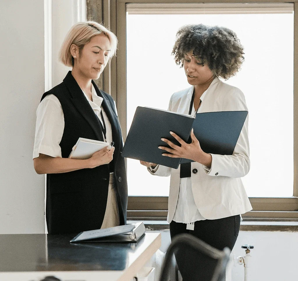 Two women in business attire reviewing documents in a modern office with a large window in the background.