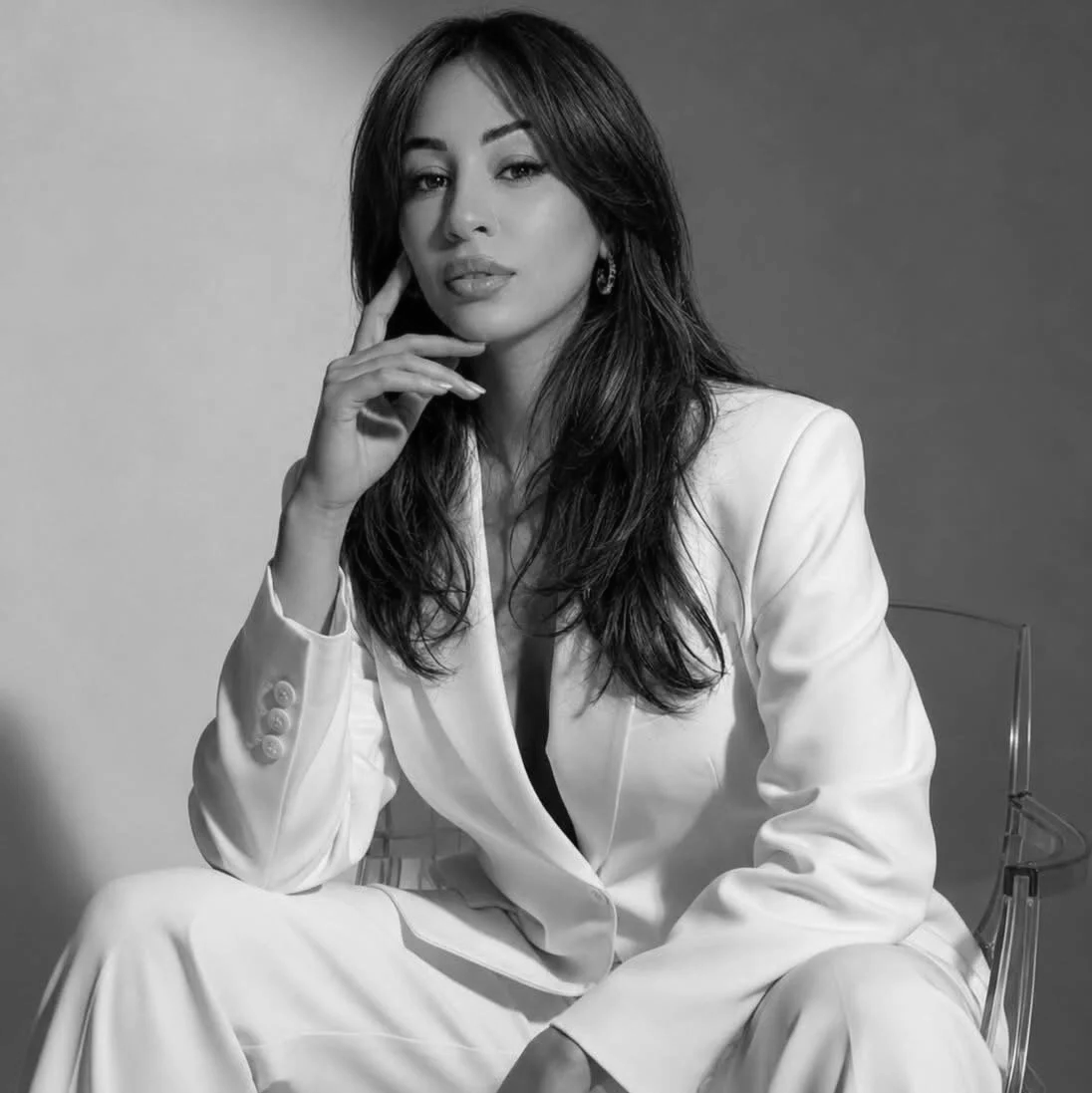 Black and white portrait of a woman with long dark hair, wearing a white blazer and pants, sitting on a transparent chair, looking at the camera with a confident expression, her hand resting on her cheek.