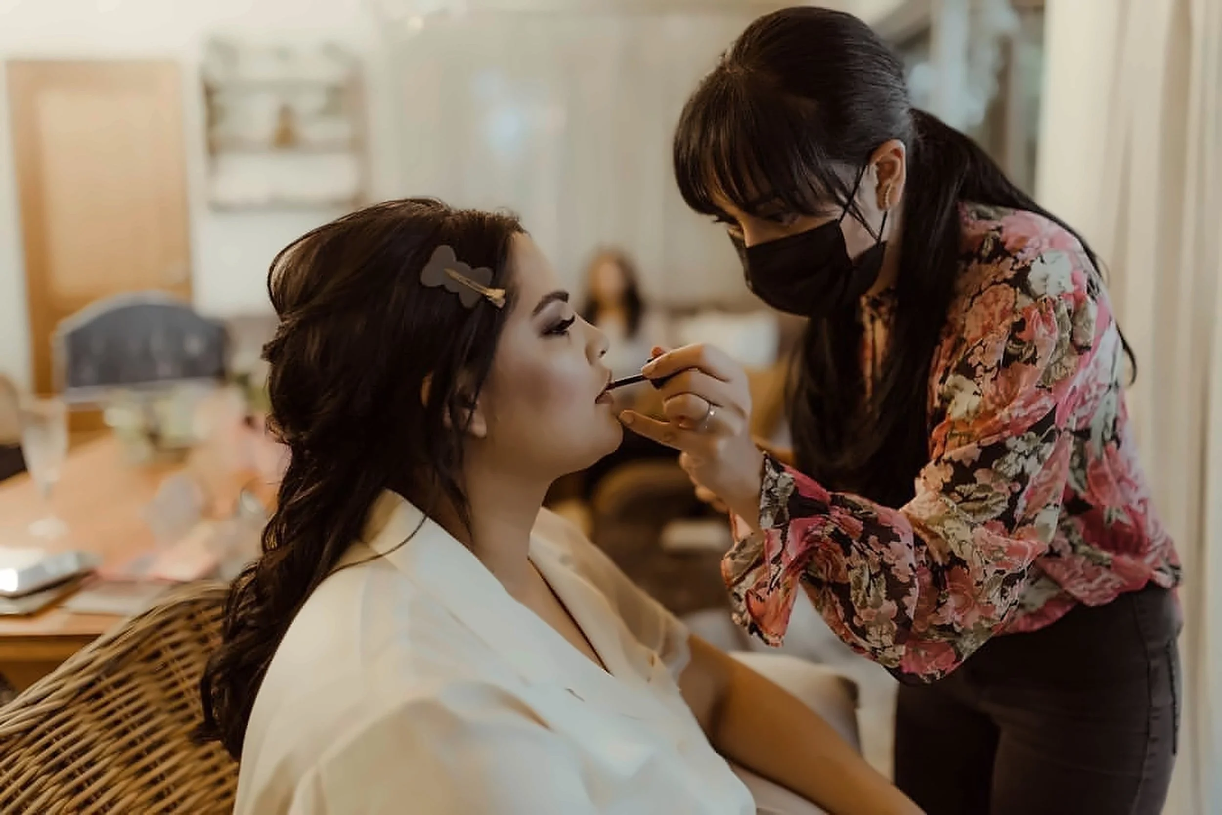A makeup artist applying lipstick to a woman seated at a vanity, with makeup brushes and beauty tools on the table, in a softly lit room.