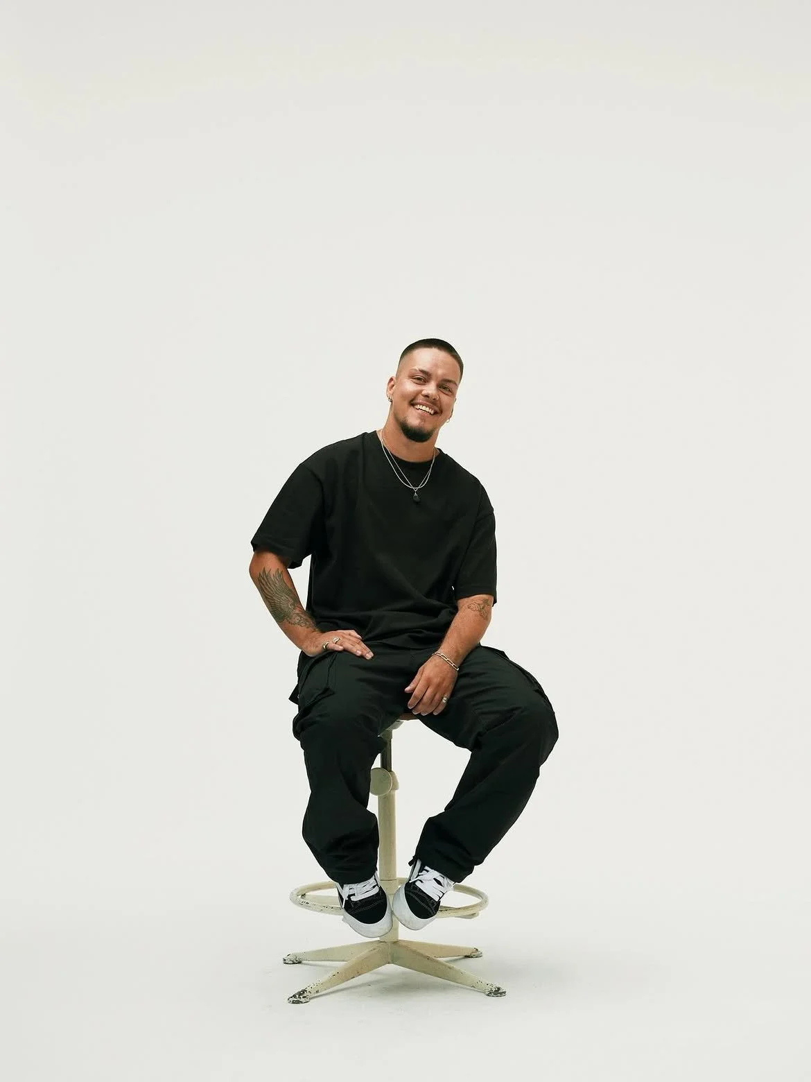 A man smiling and sitting on a stool against a plain white background, dressed in black casual clothing with tattoos on his arms.