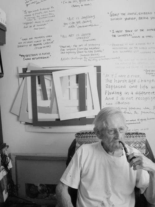 An elderly man with glasses and a beard, sitting in a room, smoking a pipe. Behind him is a wall with handwritten quotes and writings, and a framed artwork. The room appears to have a cozy, artistic atmosphere.