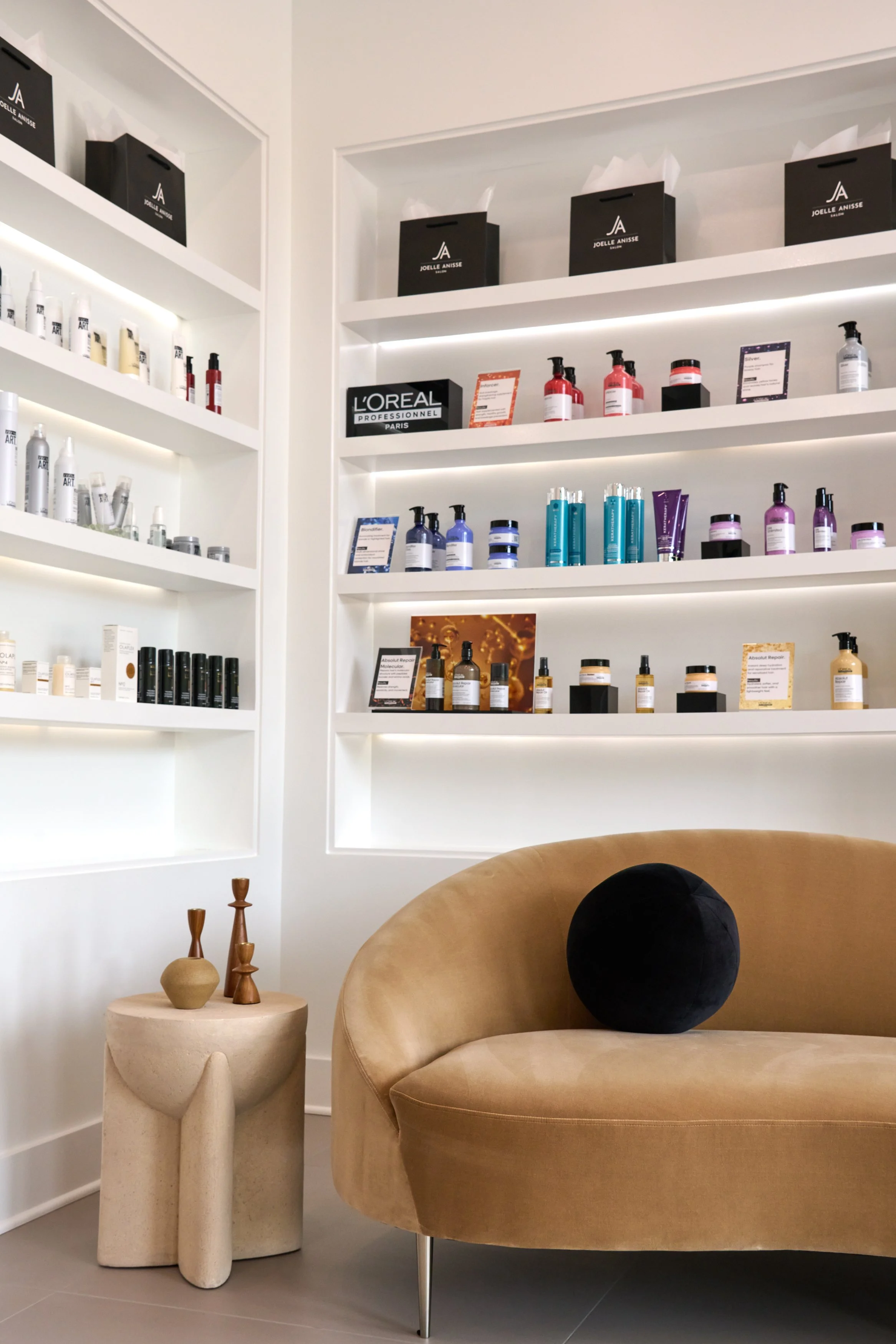 Modern interior of a beauty or skincare shop with a beige rounded sofa with a black velvet pillow, a small beige side table with decorative wooden vases, white shelves with skincare products, and black gift bags on the upper shelves.