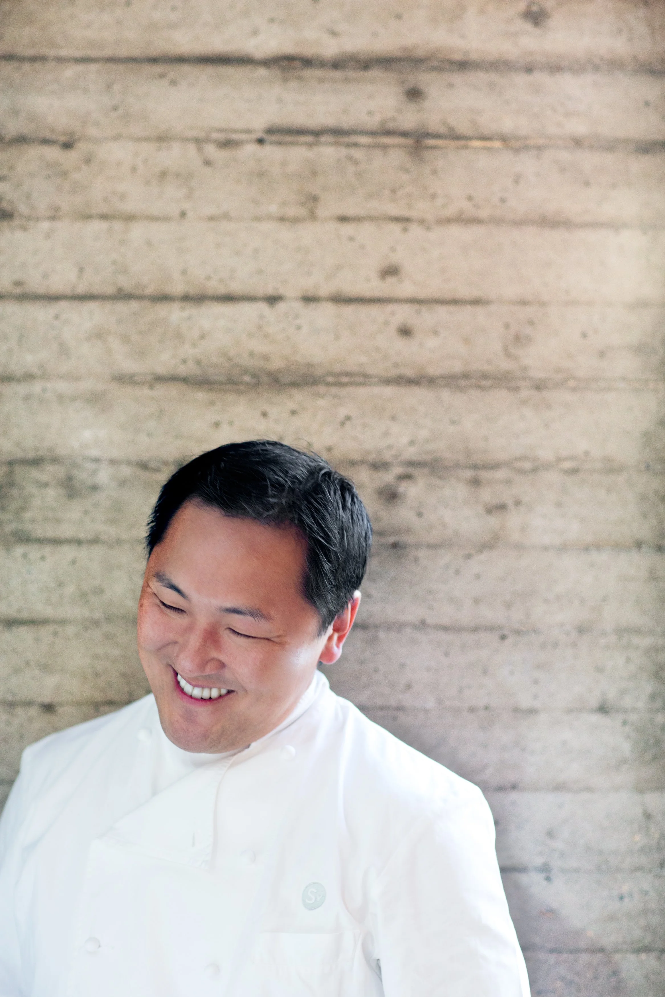 Smiling man in a white chef's coat with a wooden wall background.