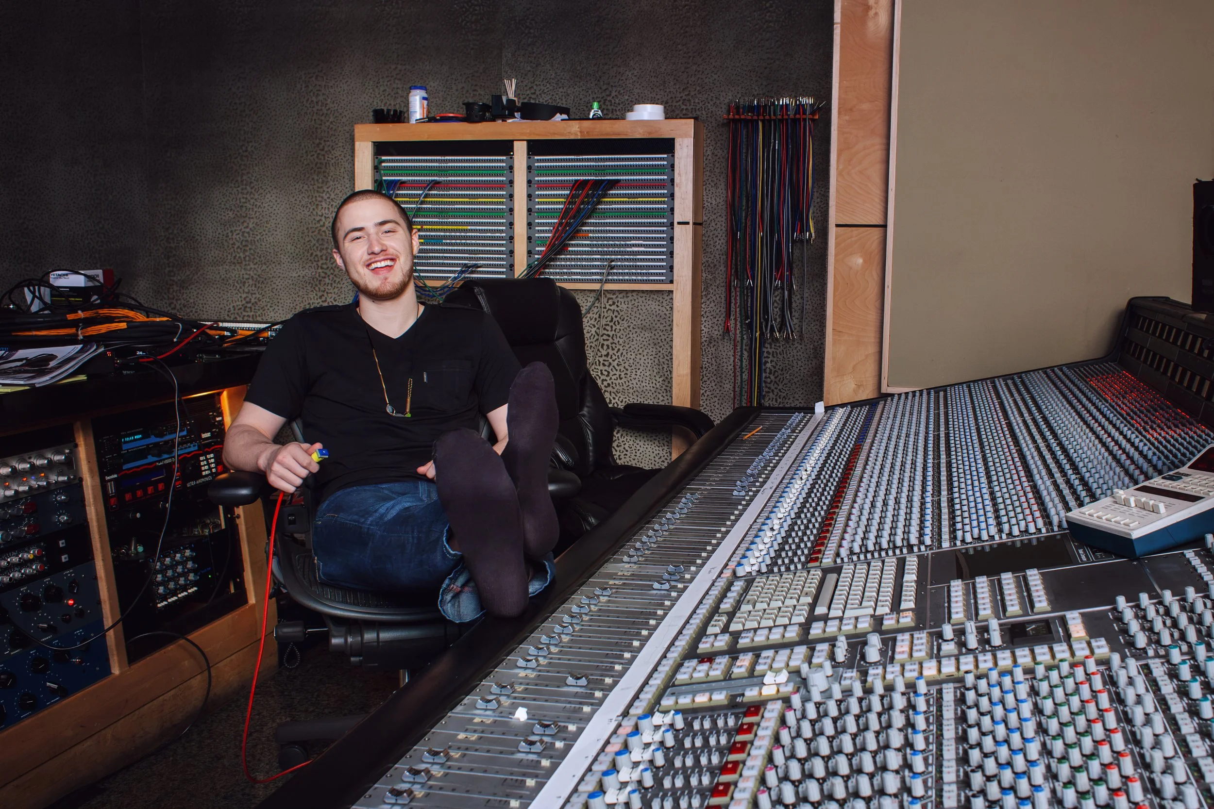 A young man with short hair and a beard sitting in front of a large audio mixing console in a recording studio, smiling and holding a yellow cable.