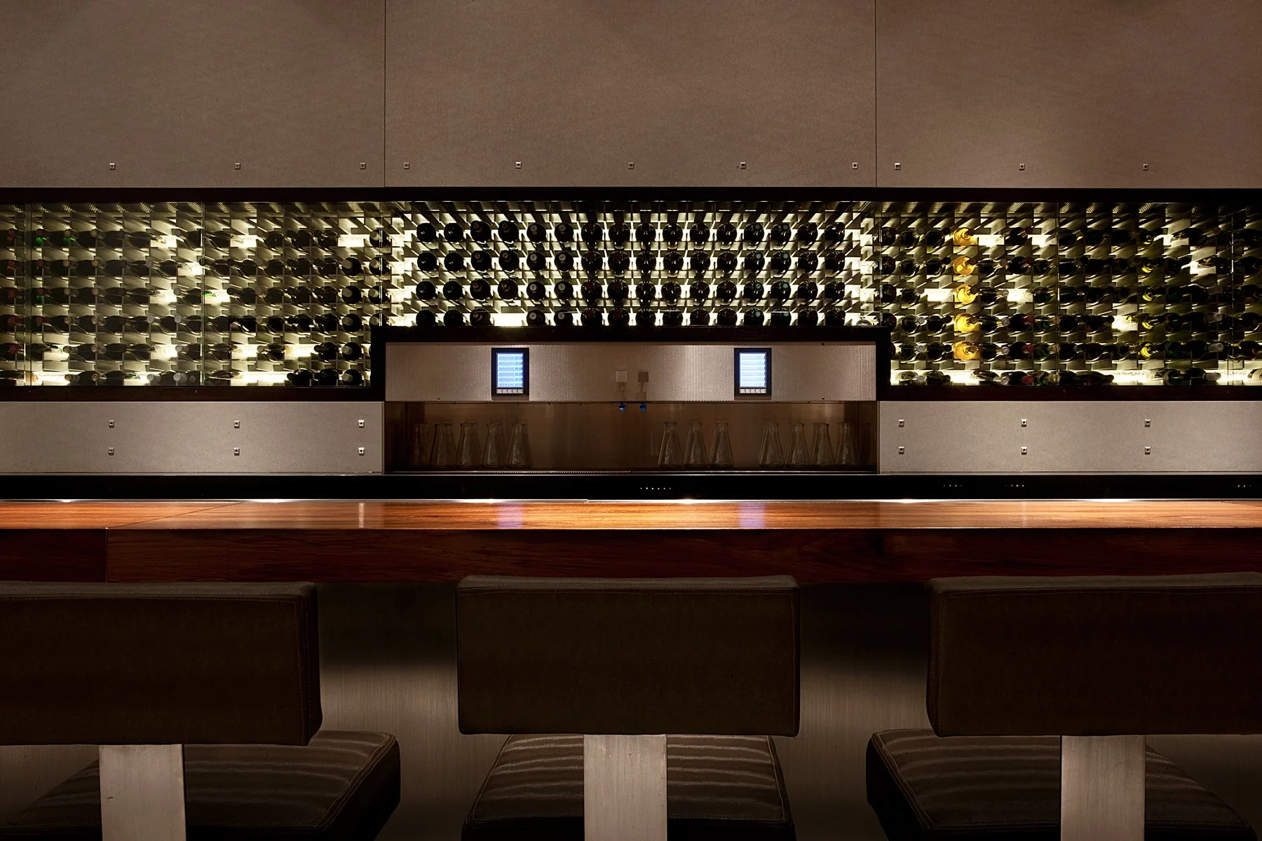 A modern wine display in a restaurant with bottles on the wall, two digital tablets in the center, and a bar counter with glasses below.