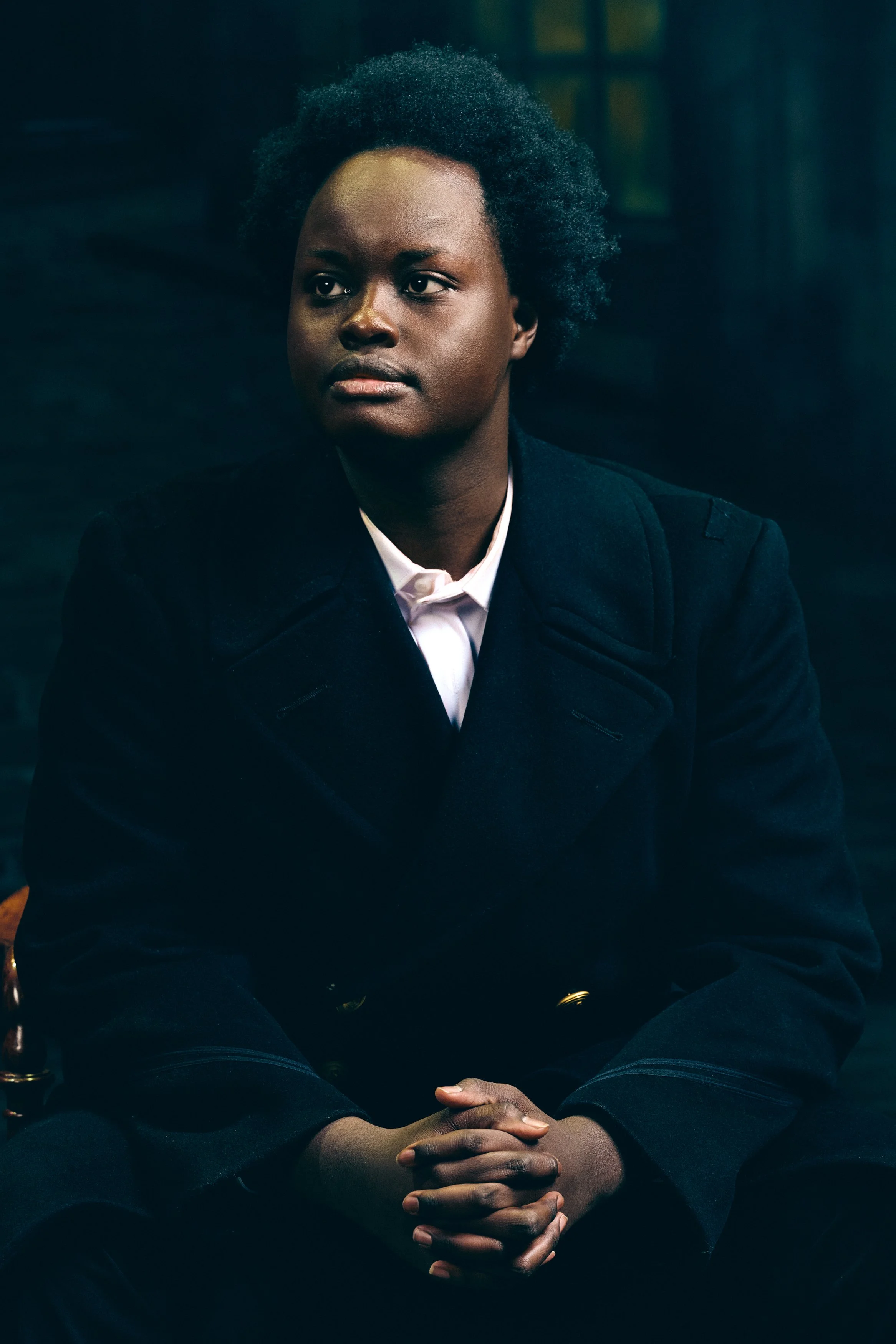 A young African American person with natural afro hair sitting with hands clasped, wearing a black coat and white shirt against a dark background.