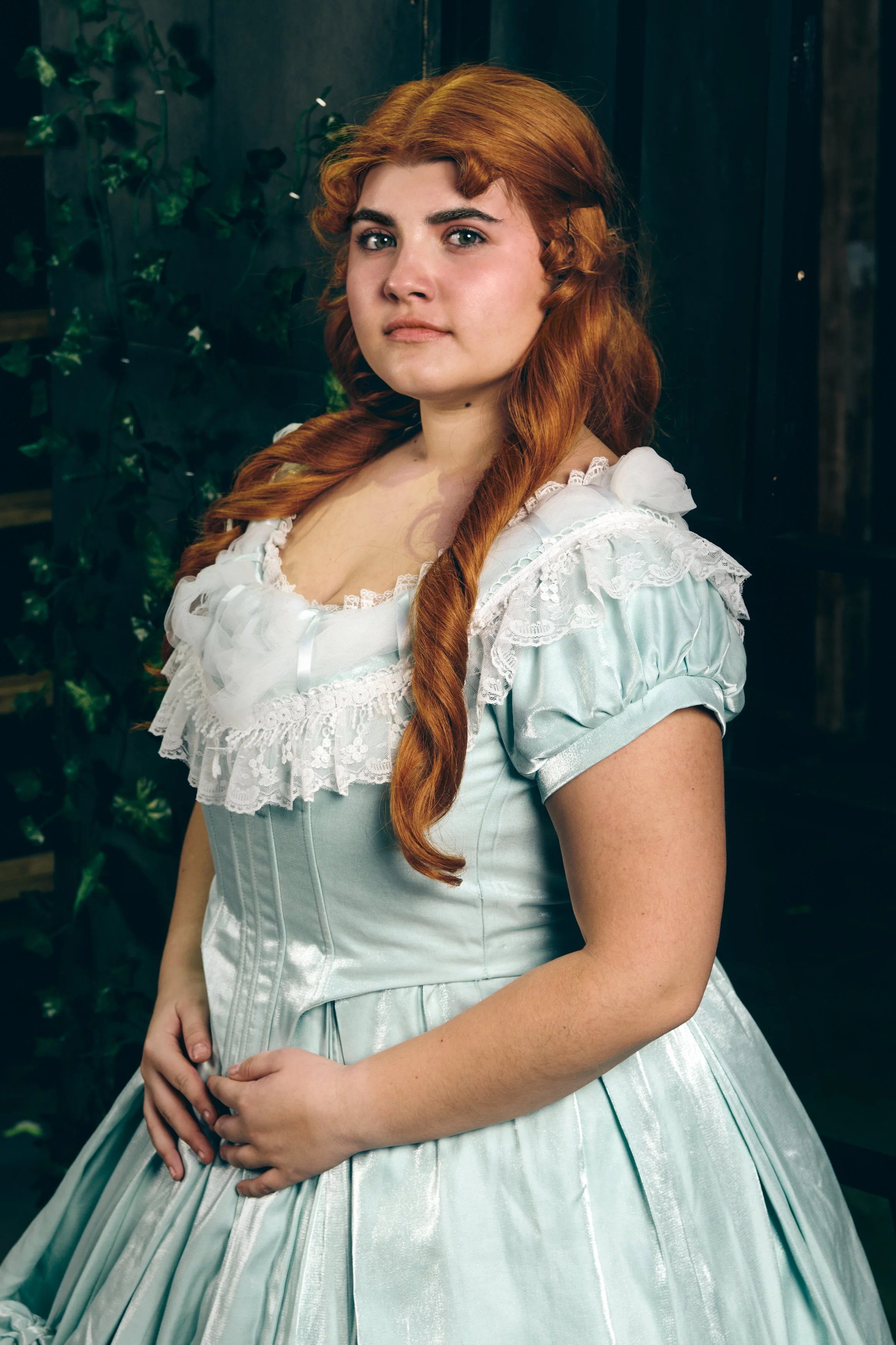 A woman with red hair wearing a light blue Victorian-style dress with lace and ruffles, standing in front of a dark background with green plants.