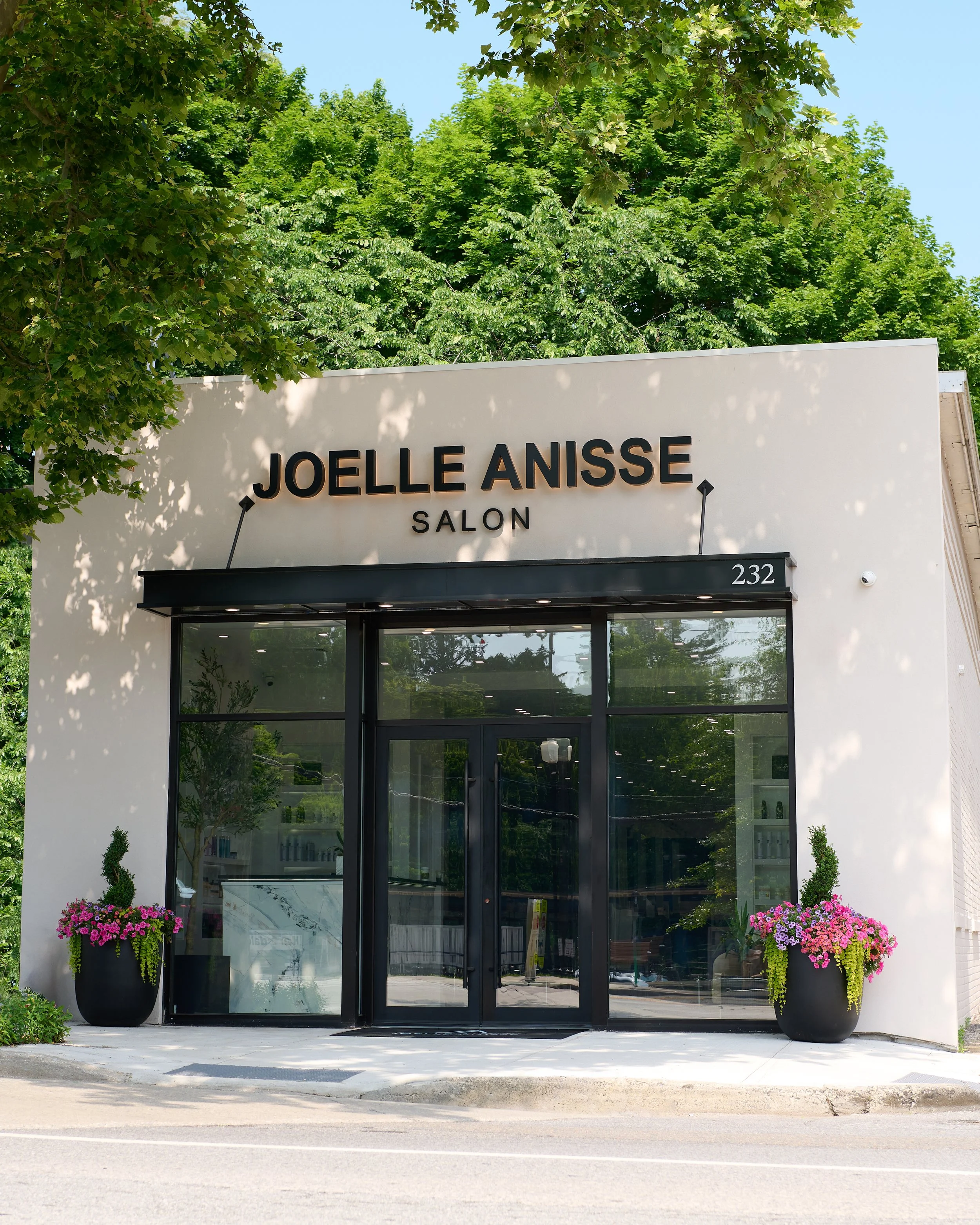 The storefront of Joelle Anisse Salon, with large black framed glass doors, two large black planters with colorful pink and purple flowers, and a white exterior wall with the salon's name and address.