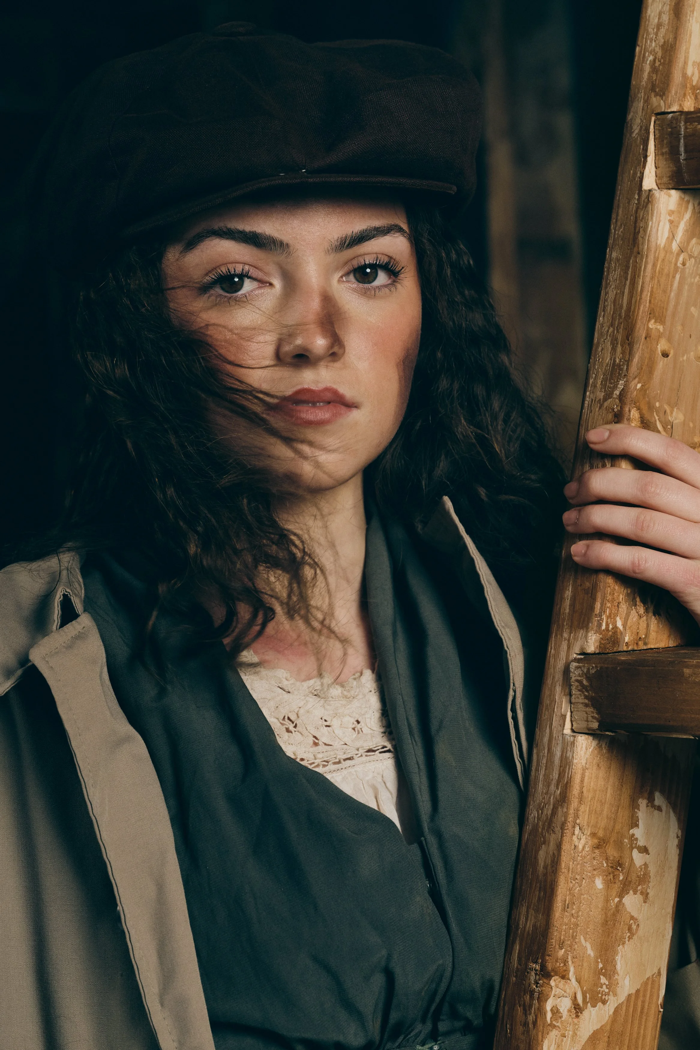A young woman with wavy dark hair, wearing a black cap and outdoor clothing, leans against a log with her hand resting on it. She has a neutral expression.