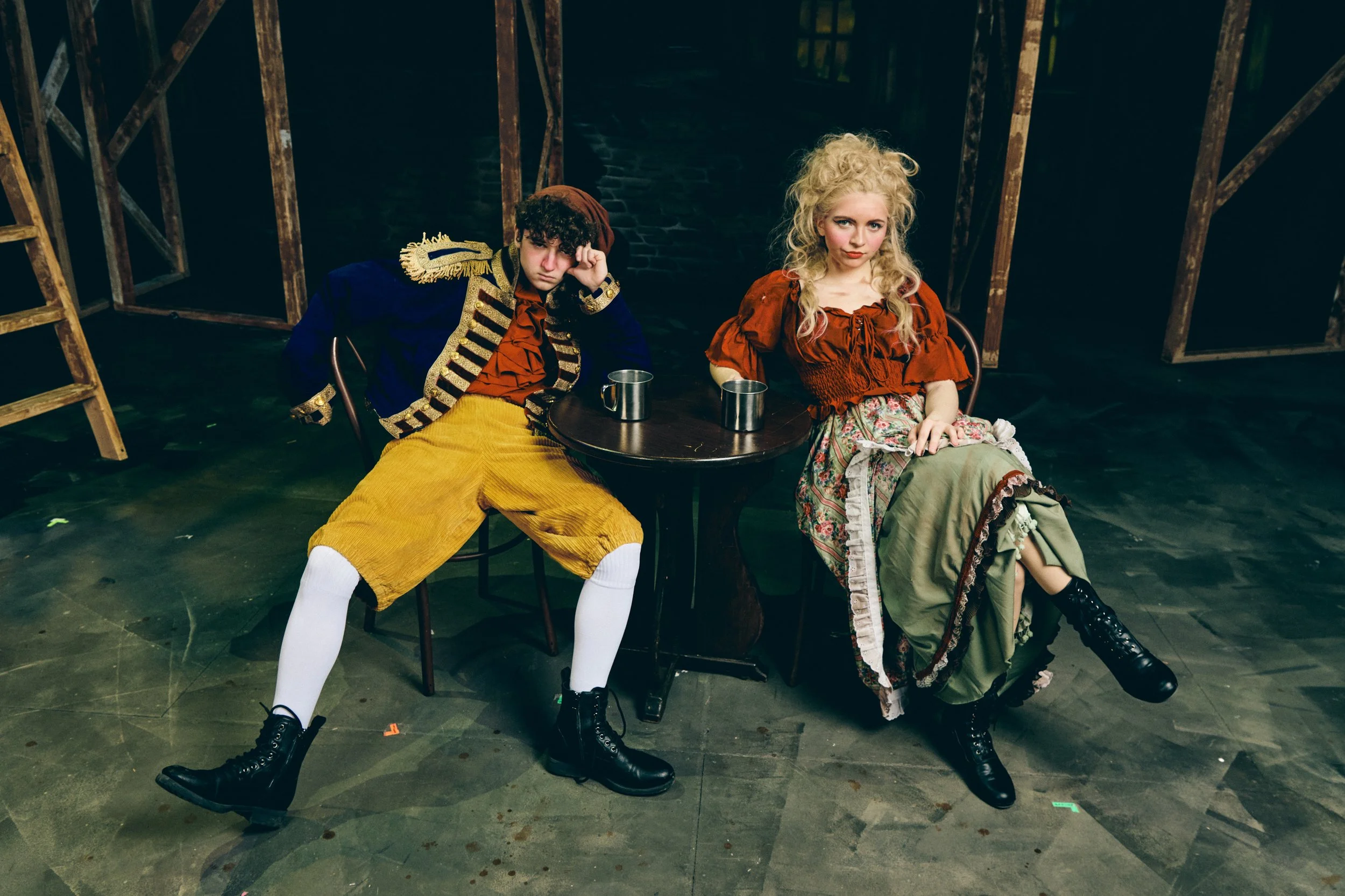 Two young women dressed in historical costumes sitting at a small round table with metal mugs, on a dark stage or set with wooden structures in the background.