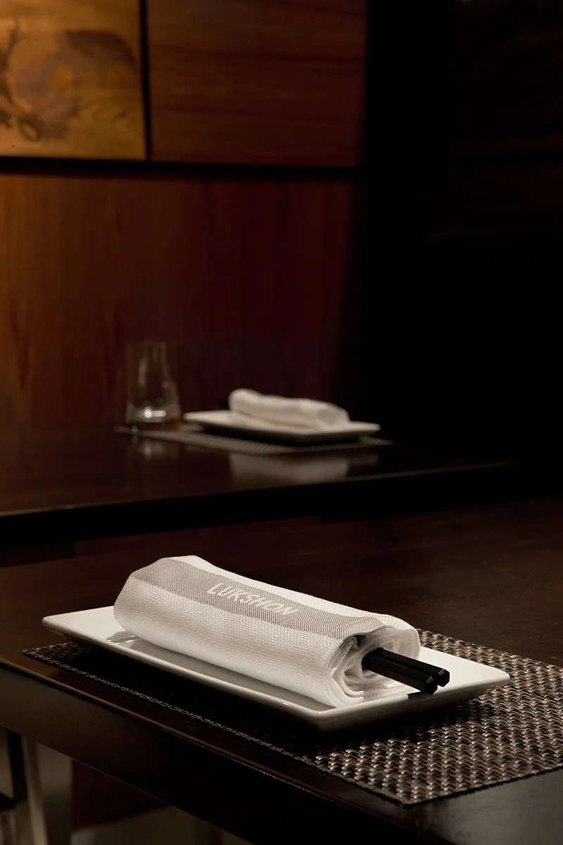 A table setting with a white plate, rolled white napkin, and black chopsticks on a woven placemat in a dimly lit restaurant.