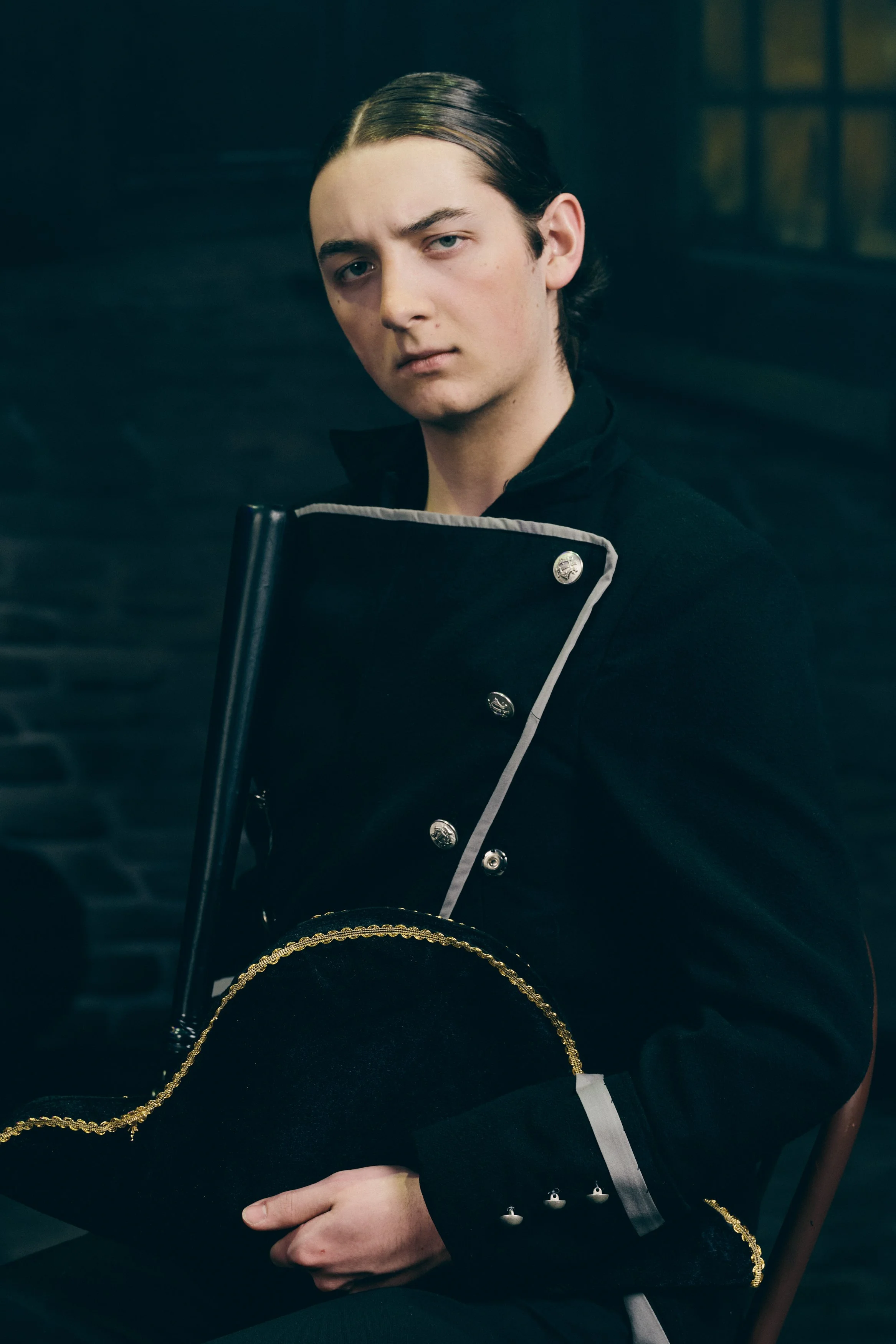 Young man with slicked-back dark hair, wearing a black military-style jacket with silver buttons, holding a black drum with gold trim, sitting in a dimly lit room.