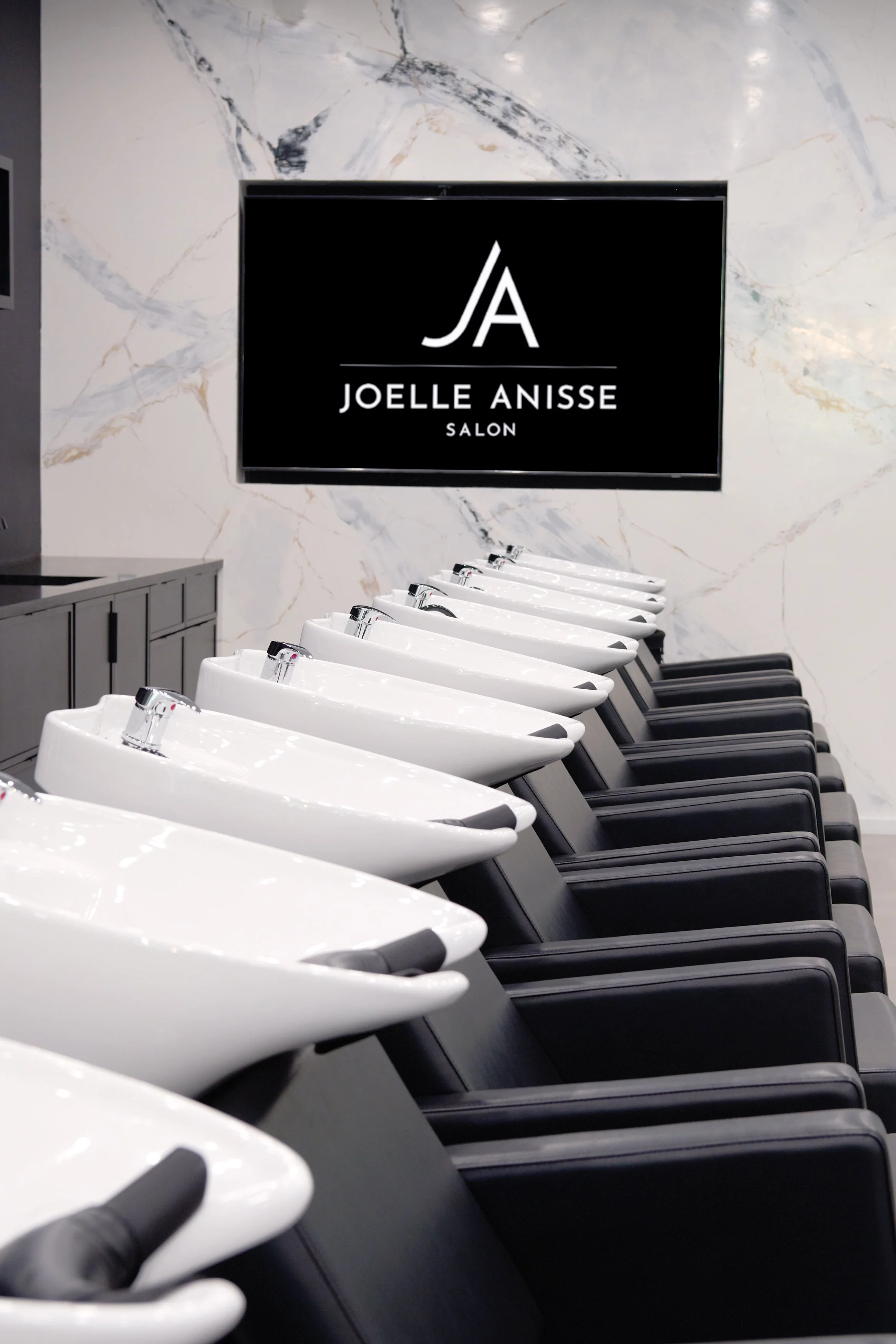 Row of white salon shampoo bowls with black chairs underneath and a black sign on the marble wall reading 'Joelle Anisse Salon' with a stylized 'JA' logo.