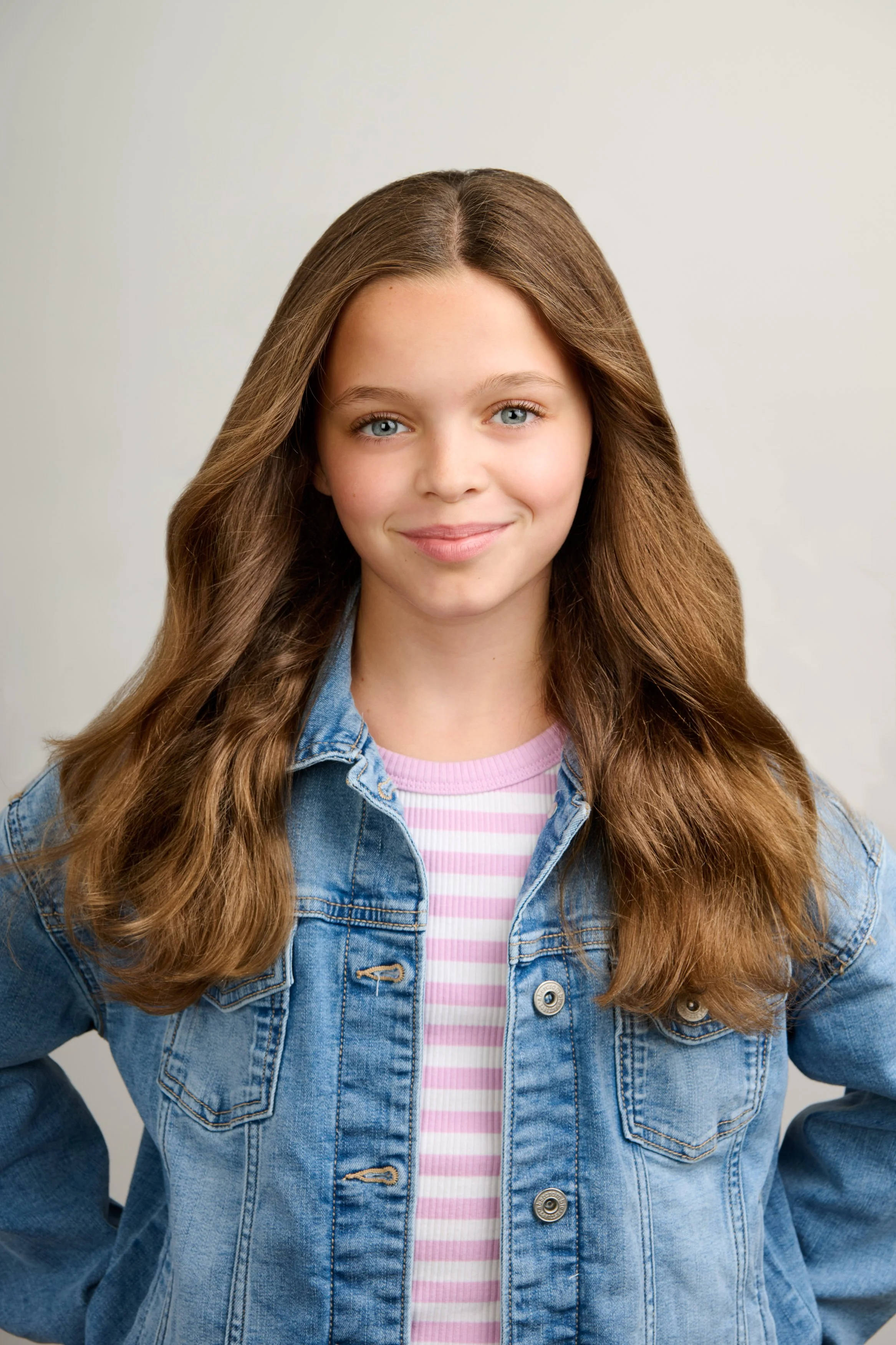 A young girl with long wavy brown hair and blue eyes smiling, wearing a denim jacket over a pink and white striped shirt, standing against a plain light-colored background.