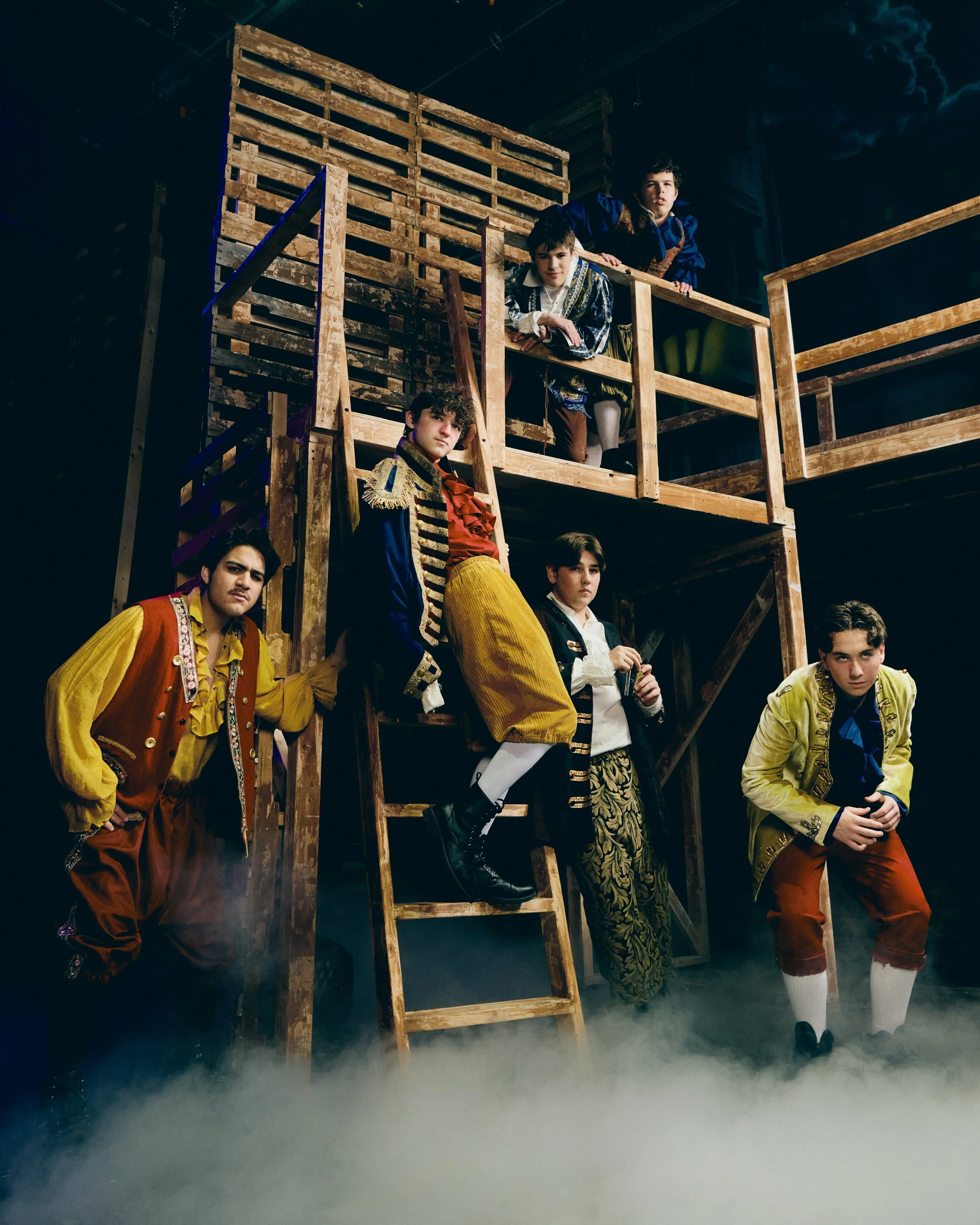 Group of young performers dressed in historical costumes, standing on a wooden stage set with a two-story balcony, against a dark, theatrical backdrop.