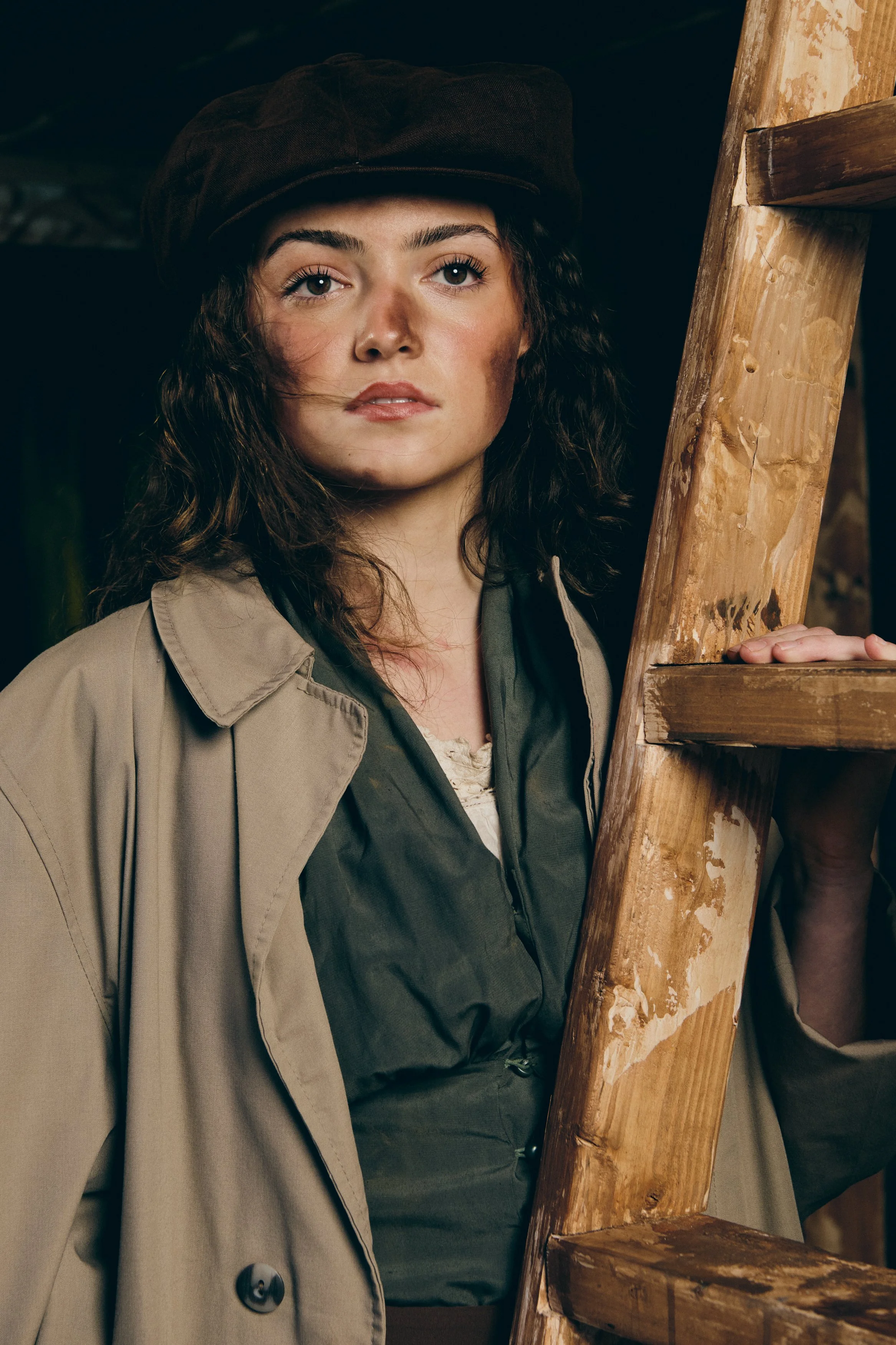 A woman with curly brown hair wearing a brown cap, a beige trench coat, and green clothing stands near a wooden ladder, looking at the camera with a neutral expression.