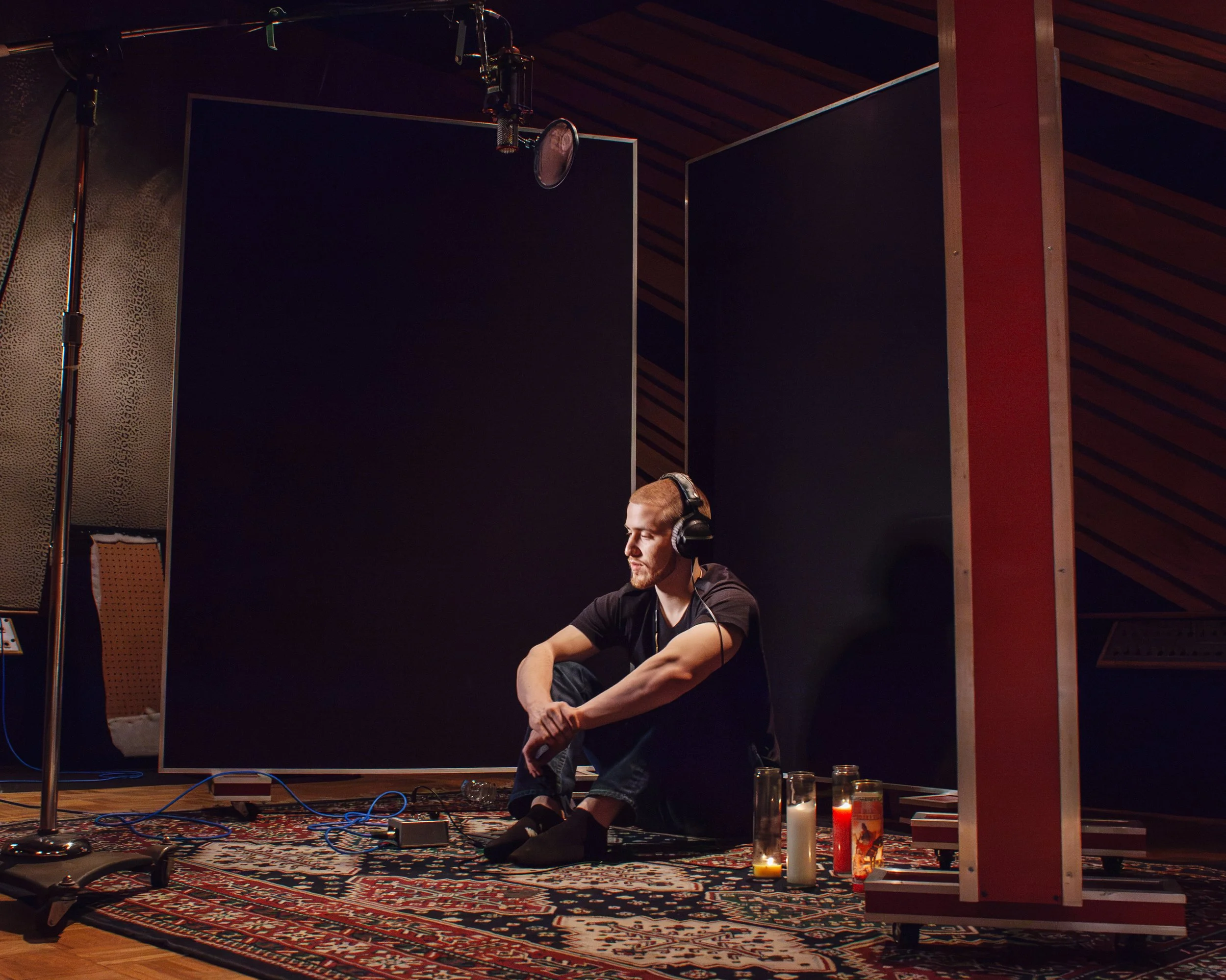 A man with headphones sitting cross-legged on a patterned rug in a recording studio with dark walls and wooden ceiling. Several candles and bottles are placed in front of him, and recording equipment, including a microphone, is visible above him.