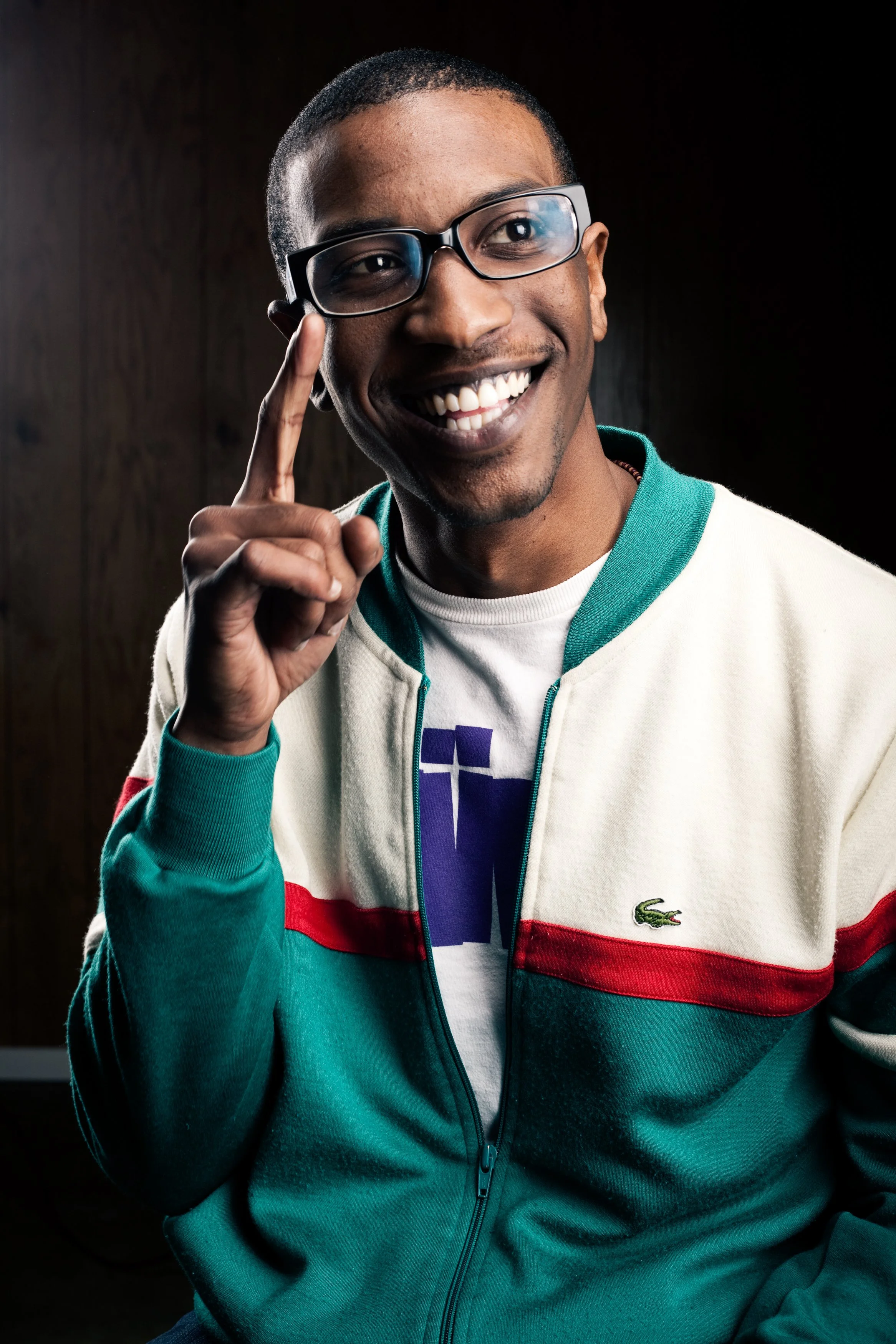A smiling man with glasses, short hair, wearing a colorful Lacoste jacket, pointing his index finger to his temple, against a dark background.