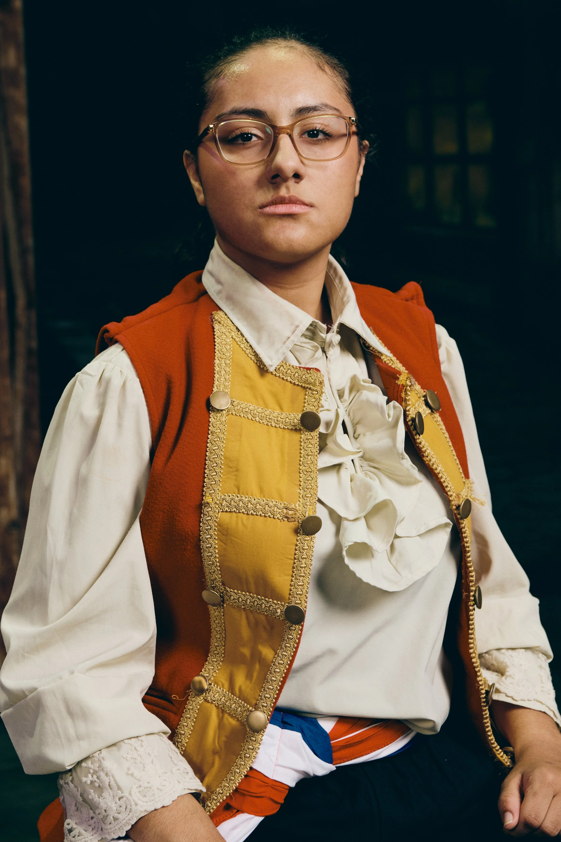 A woman wearing glasses, a white blouse with ruffle detail, a red and yellow embroidered vest, and lace trim on her sleeves, with short pants featuring a color pattern, sitting against a dark background, looking directly at the camera with a serious 