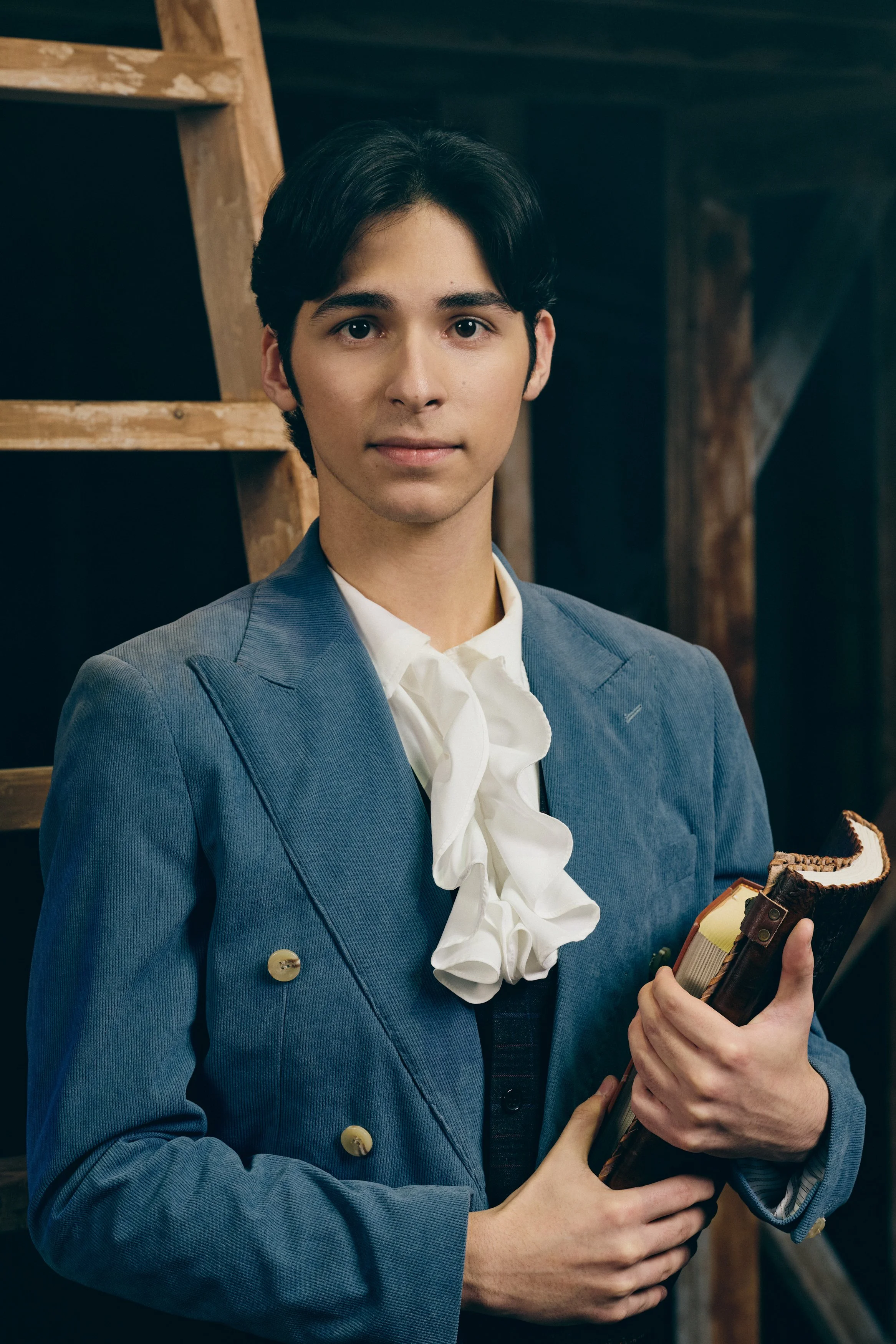 Close-up of a young man with dark hair wearing a blue blazer and holding books, standing indoors near wooden shelving.